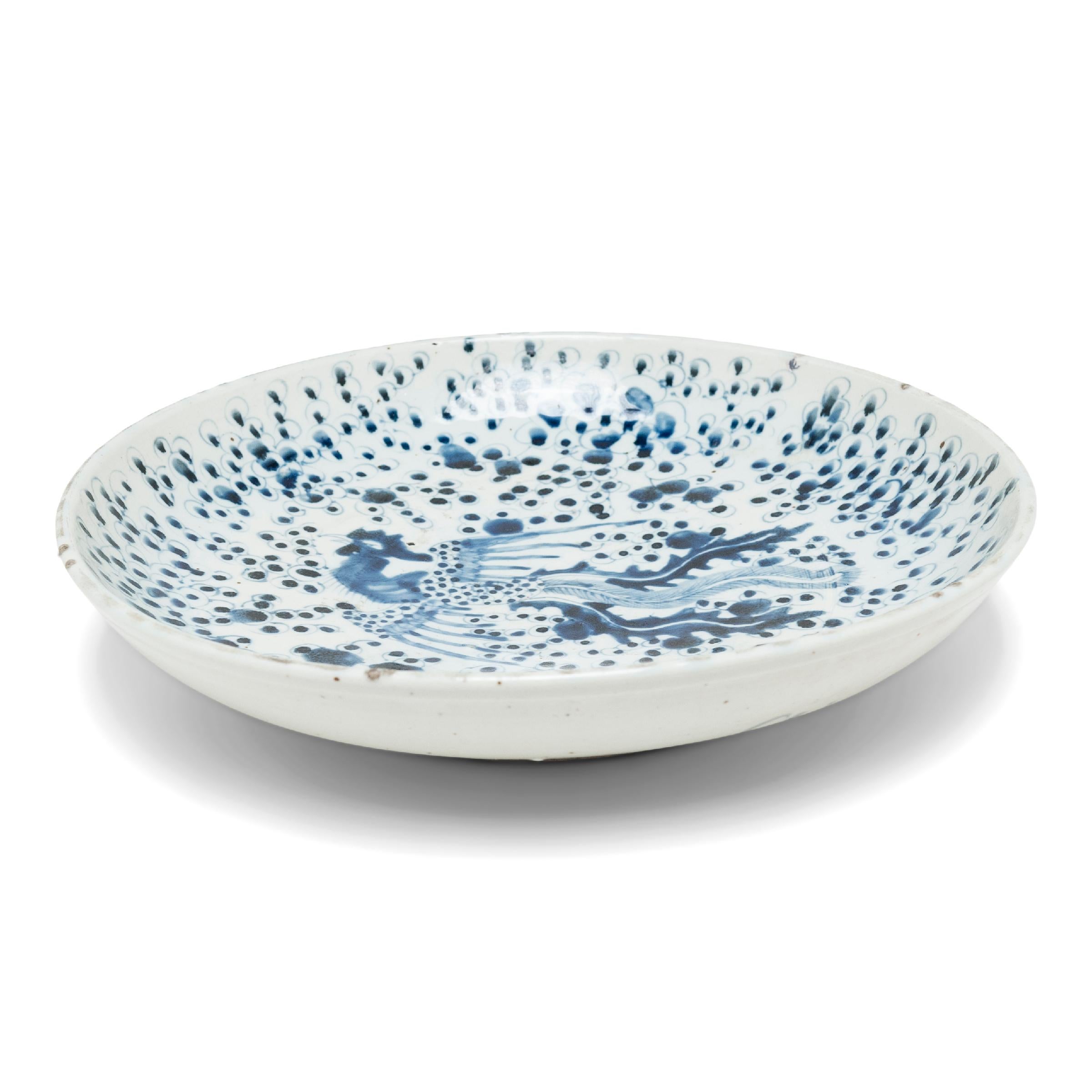 An elevated example of Chinese blue-and-white porcelain, this large footed dish charms with dark, cobalt-blue brushwork atop a cool white field. The plate is decorated with an all-over fish-roe pattern, resembling clouds swirling around the central