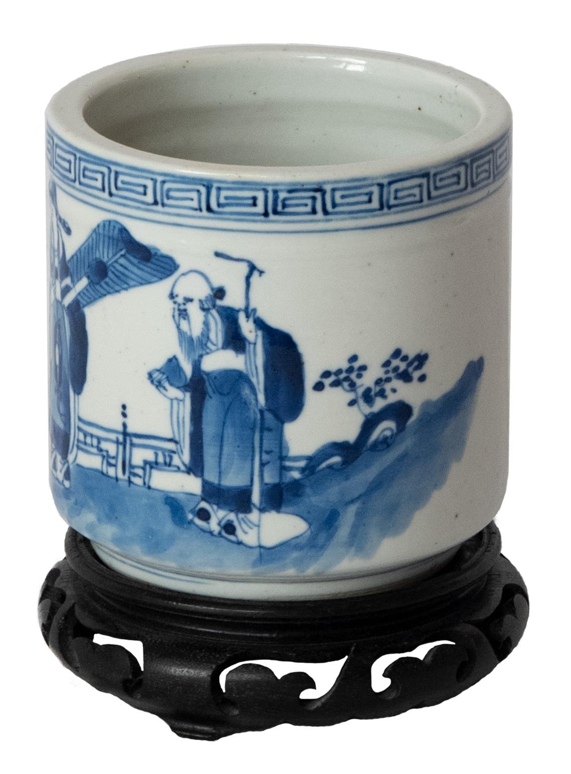 This blue and white porcelain brush pot was intended to hold brushes, pens, and other calligraphy tools. Brushed with a rich blue glaze, the pot is painted with four figures in a rural landscape. En suite with a shaped and scroll-cut hardwood stand.