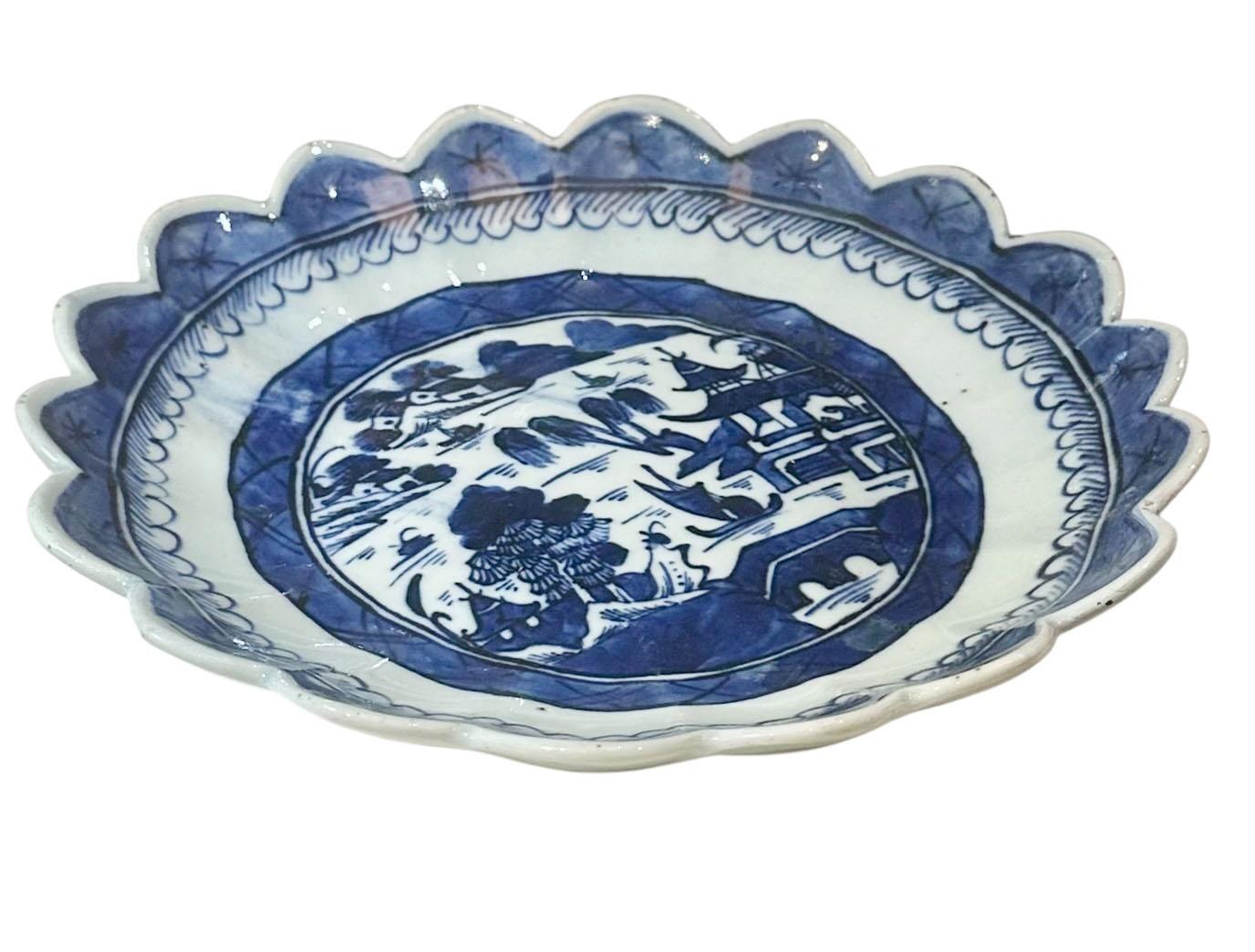 A refined early 19th century Chinese blue and white porcelain plate featuring a beautifully detailed village scene set along the water. The landscape is delicately hand-painted in deep cobalt tones, characteristic of the finest export porcelain of