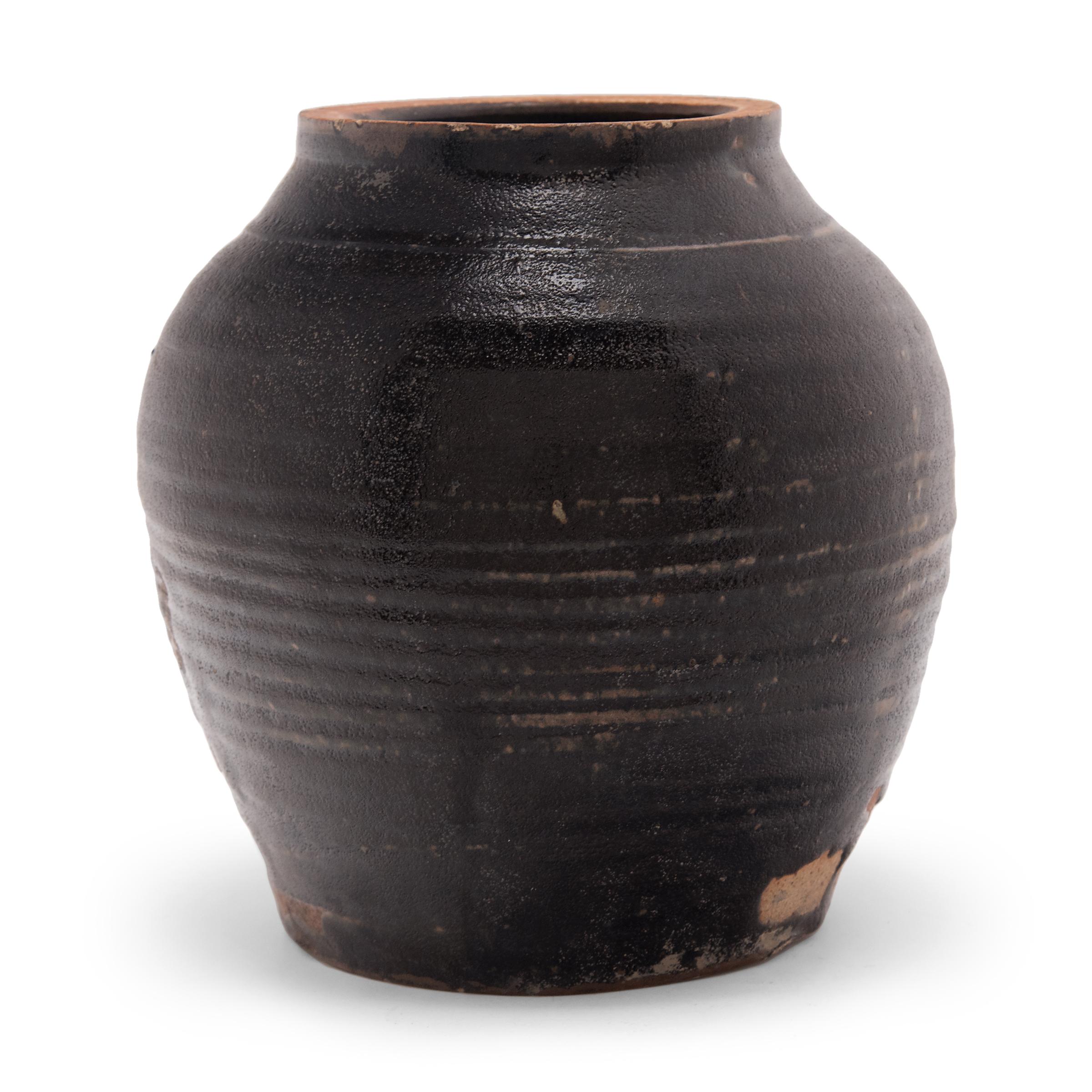 This early 20th century jar is coated in a thick brown glaze that spreads unevenly across the ridged sides, revealing areas of the stoneware body underneath. As evidenced by the interior glaze, the jar was once used to store wet foods in a