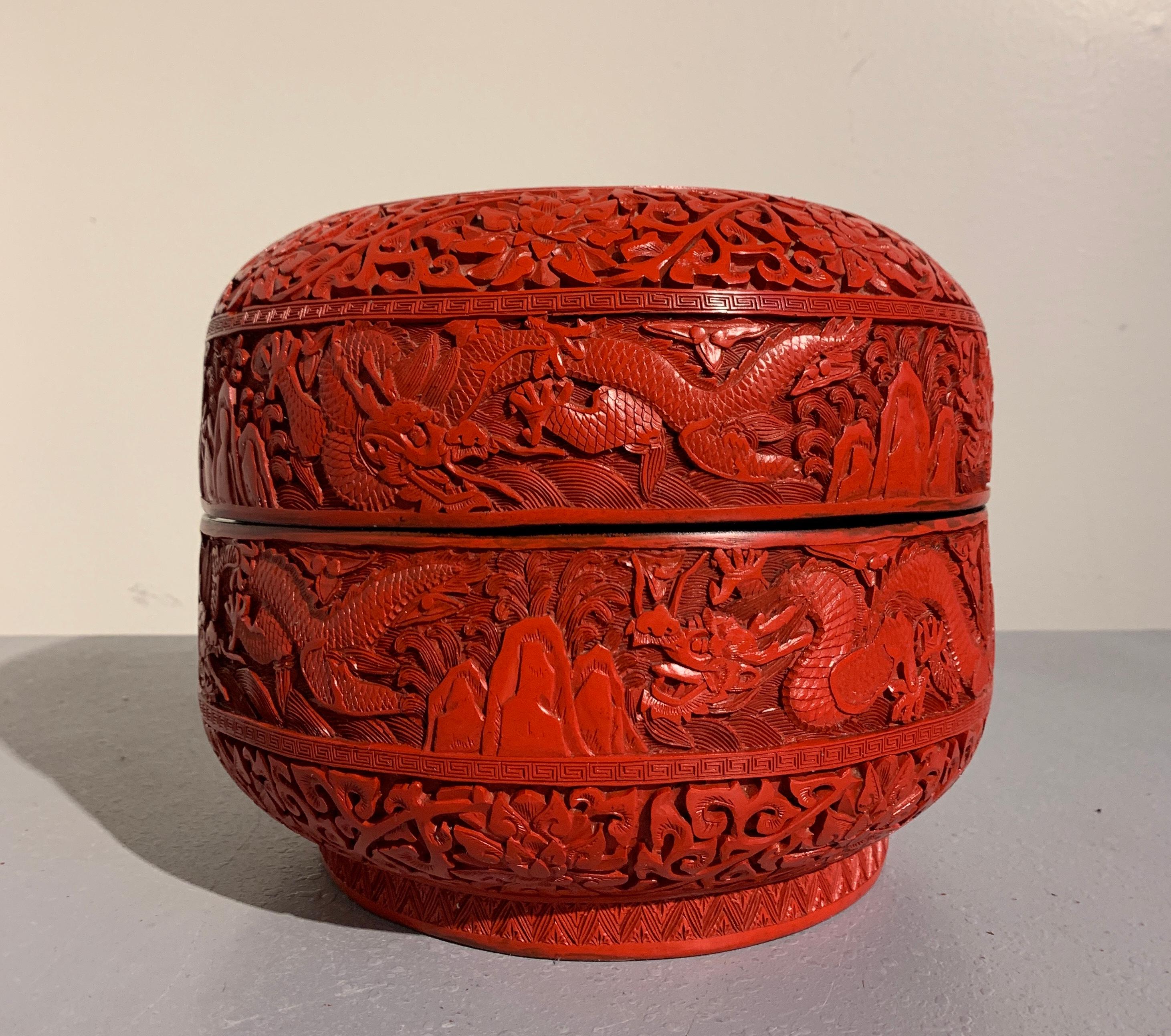 A large Chinese carved red cinnabar lacquer nine dragon cake box, late Republic period, circa 1940.
Exuberantly carved in deep relief with a design nine five clawed dragons dancing above rocky peaks amongst the waves and clouds, the tall, round box