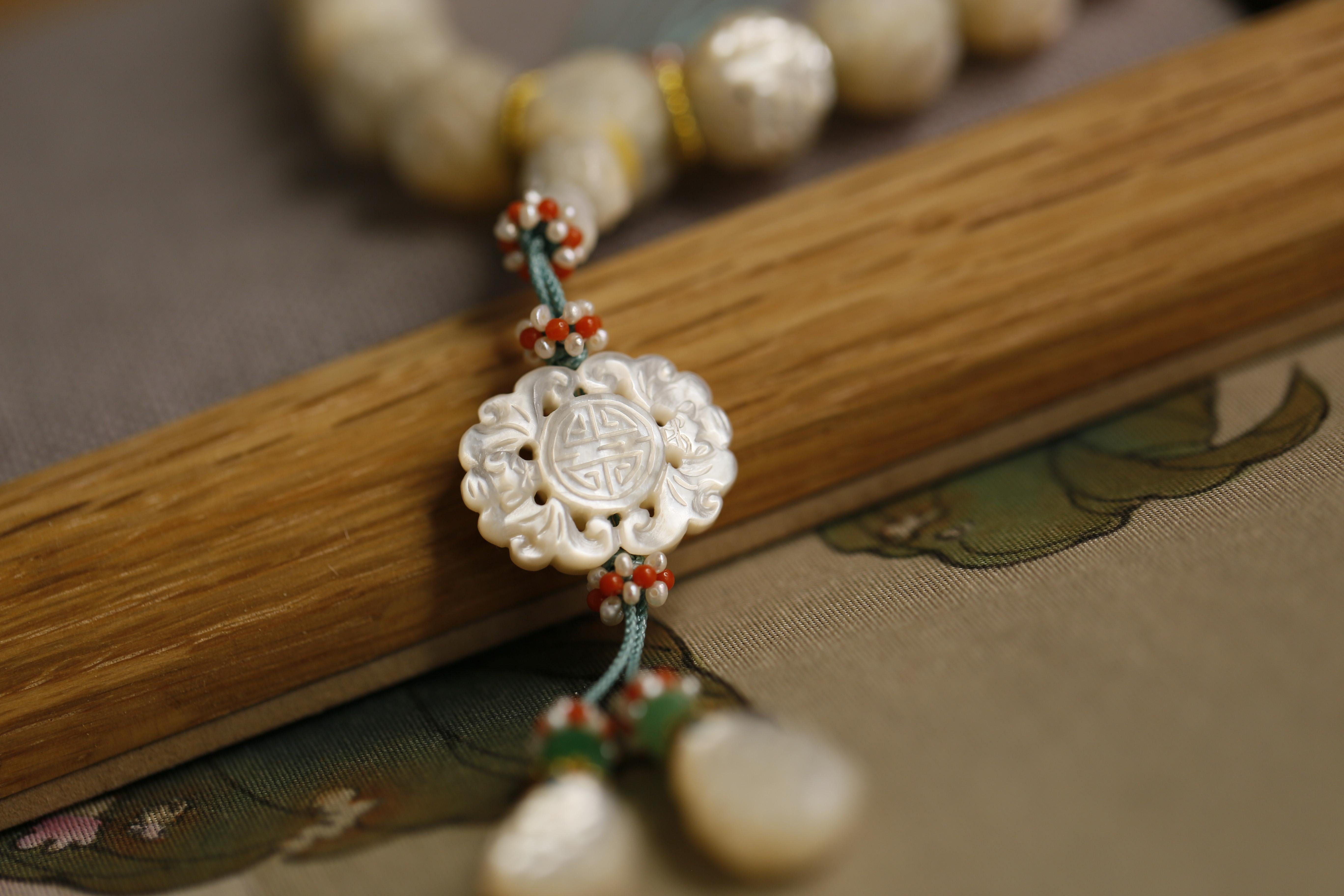 This bead bracelet is made up of carved oyster shell beads in the classic Chinese style. These beads are interspersed with green jade beads carved in the same style. Hanging from the main string is a carved plaque pendant. 