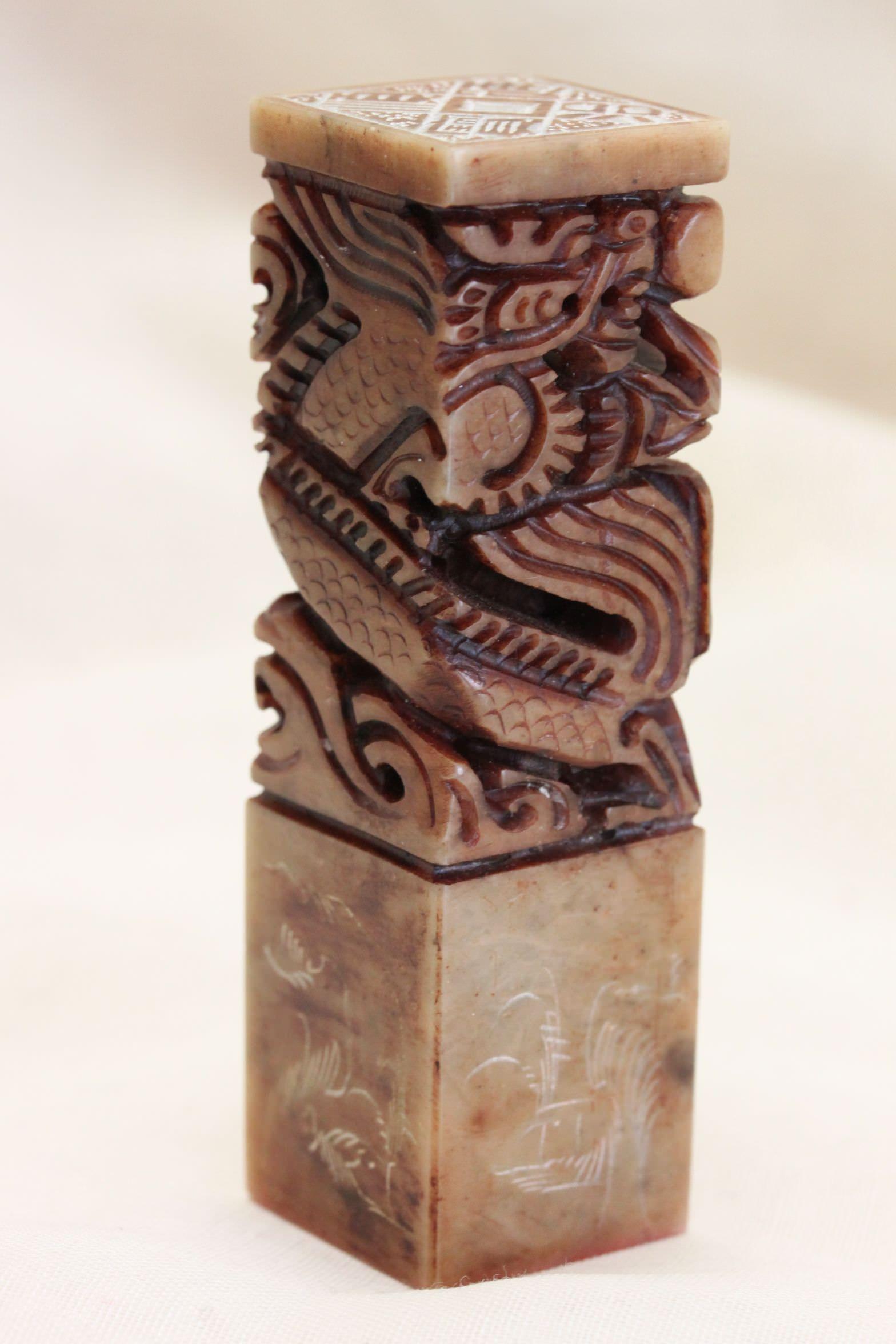 This Chinese carved soapstone seal features carving on all sides. The main carving is of a dragon, deeply cut and encircling the piece. Below the dragon are feather or fern like decorations and some Chinese characters in a circle. The top is carved