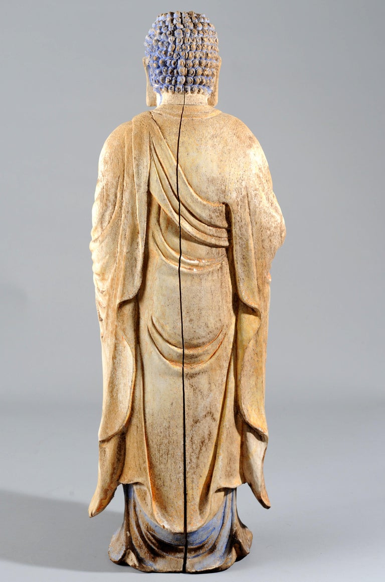 A statue of standing Buddha (Shakyamuni), finely carved out a single block of wood (might be cypress based on the wood grains). Attired in a flowing robe with open chest, the Buddha is depicted standing with both of his hands in Vitarka Mudra. The