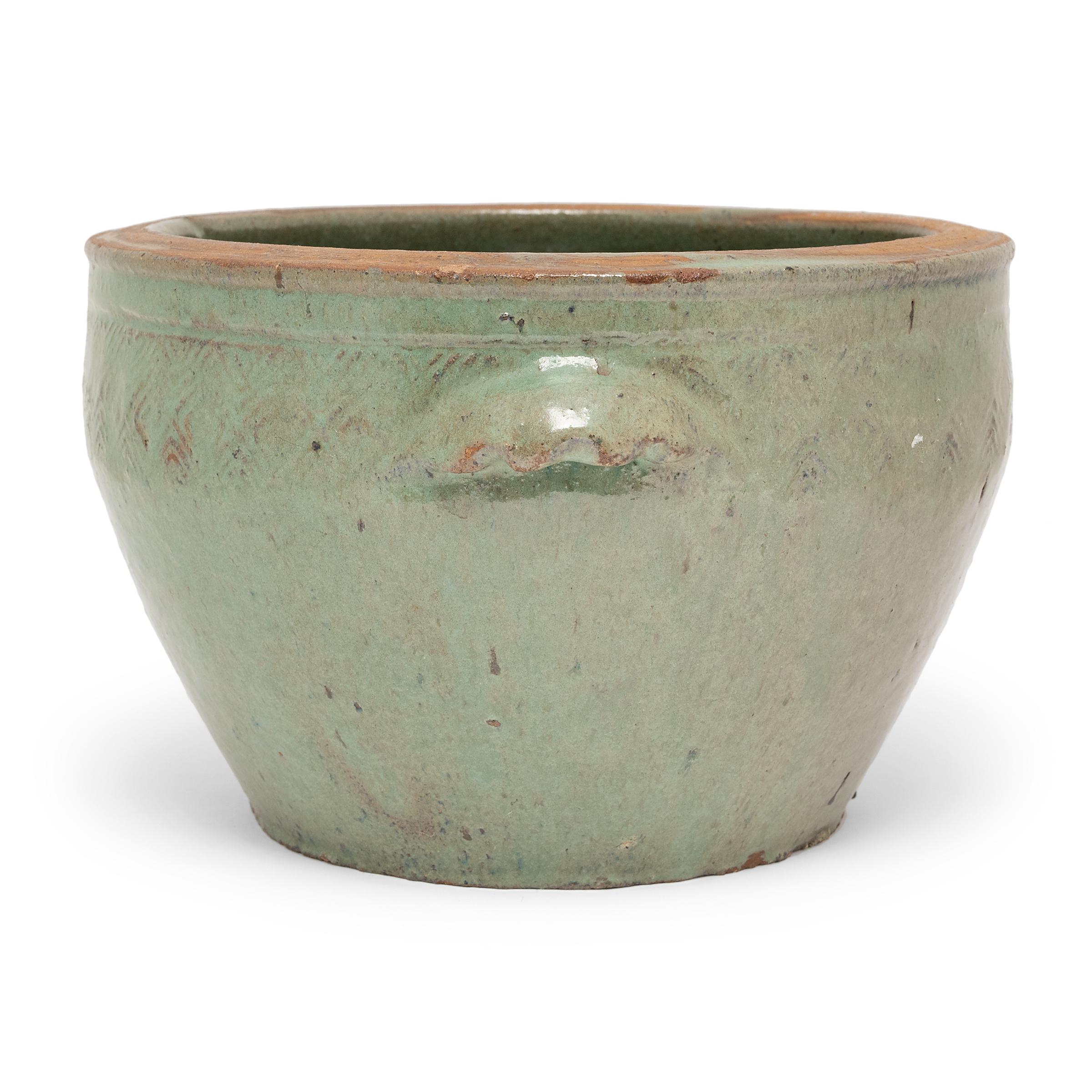 This low ceramic pot is glazed inside and out with a pale celadon-green glaze that lingers beautifully on every imperfection. The basin has a tapered form, with a wide mouth, rounded rim, and two simple side handles. A subtle diamond pattern