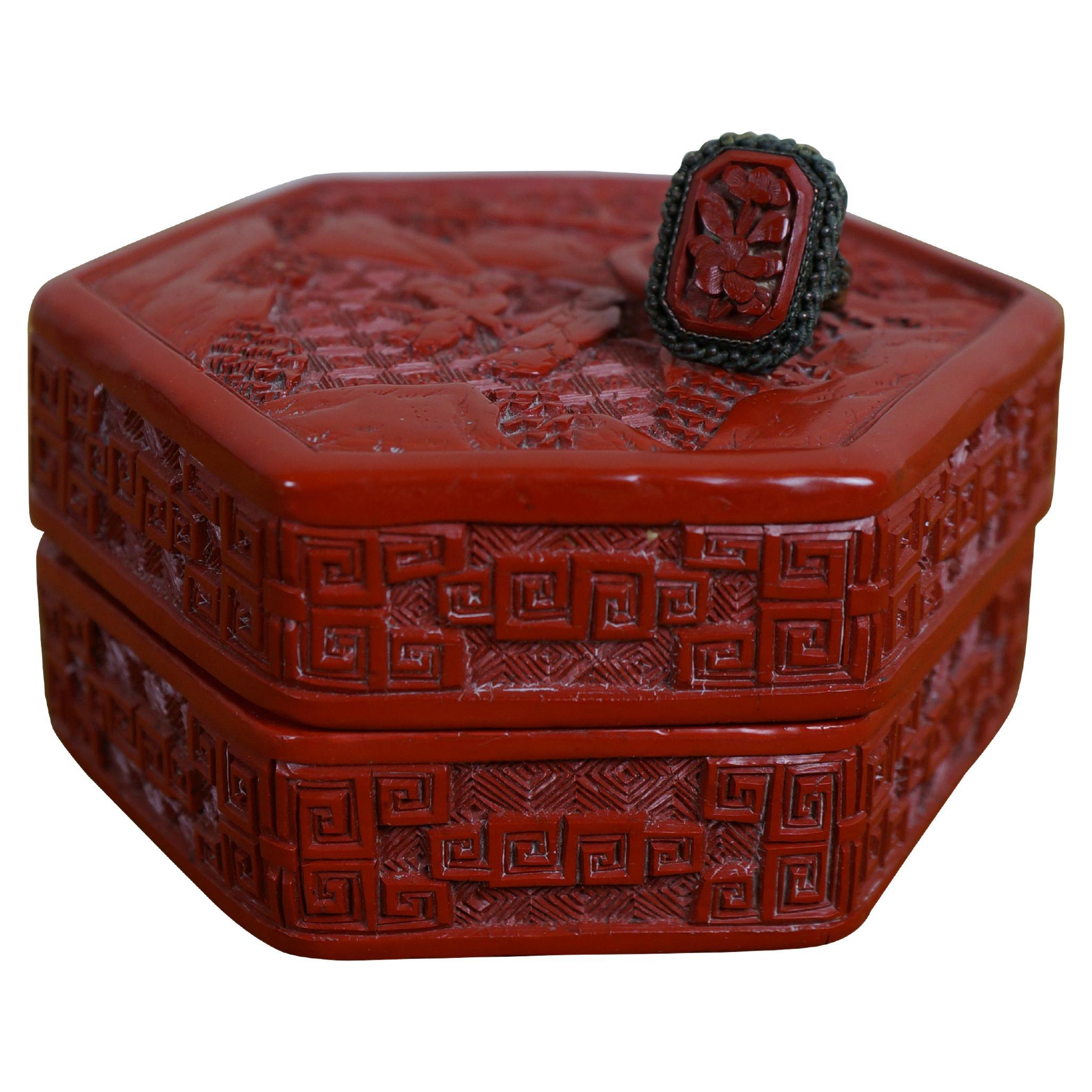 Caja de baratijas vintage de cinabrio rojo del siglo XX con anilla. Caja de forma hexagonal con motivos geométricos alrededor de los lados y la tapa, en la que aparece una pareja de figuras bajo un pino nudoso. El anillo presenta un motivo floral