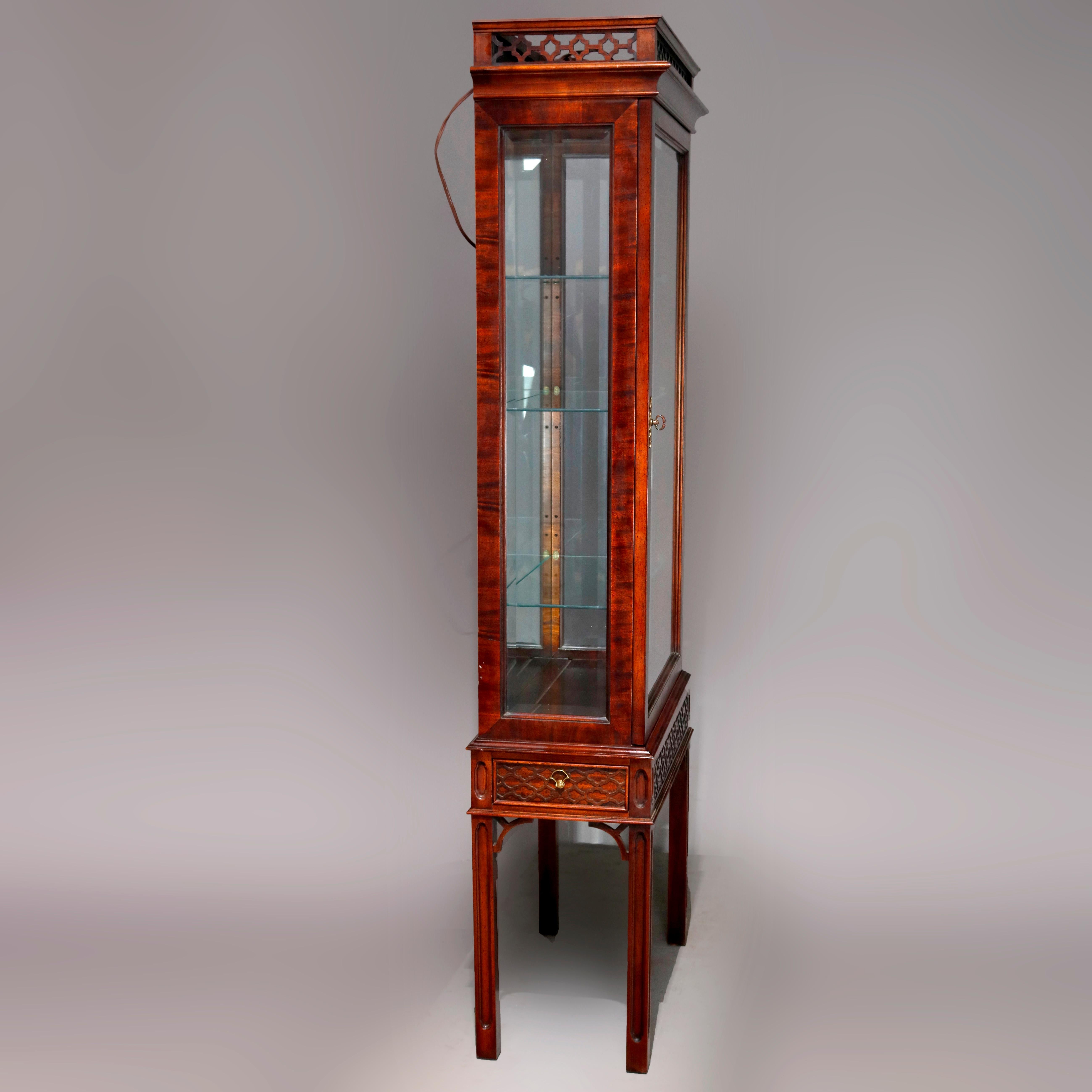 A Chinese Chippendale curio vitrine offers flame mahogany construction with carved gallery surmounting lighted and mirrored curio case with single door, shelved interior, and carved skirt, raised on straight and square legs with cutout corbels, 20th