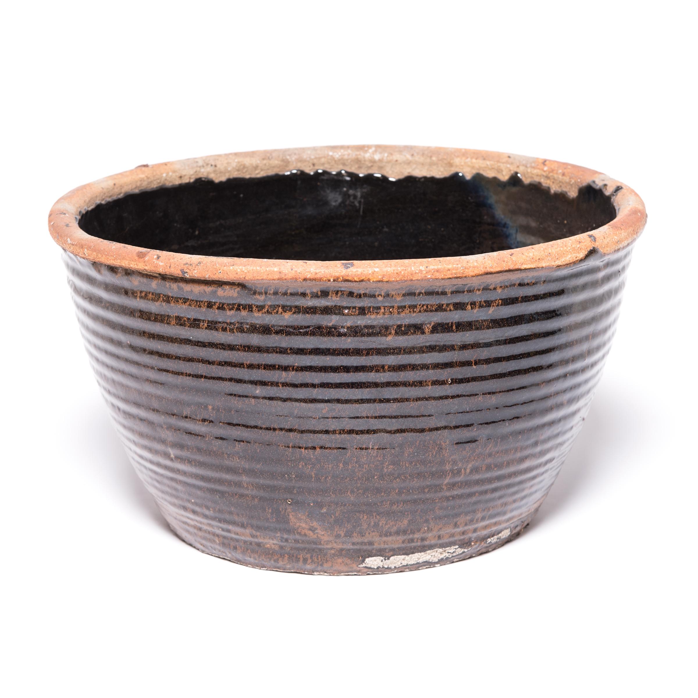 Originally used for pickling foods, this mid-20th century ceramic jar is coated inside and out with a dark glaze. Subtle ridges suggest that the bowl was fashioned by coiling flattened ropes of clay into the desired shape. Featuring an unglazed rim