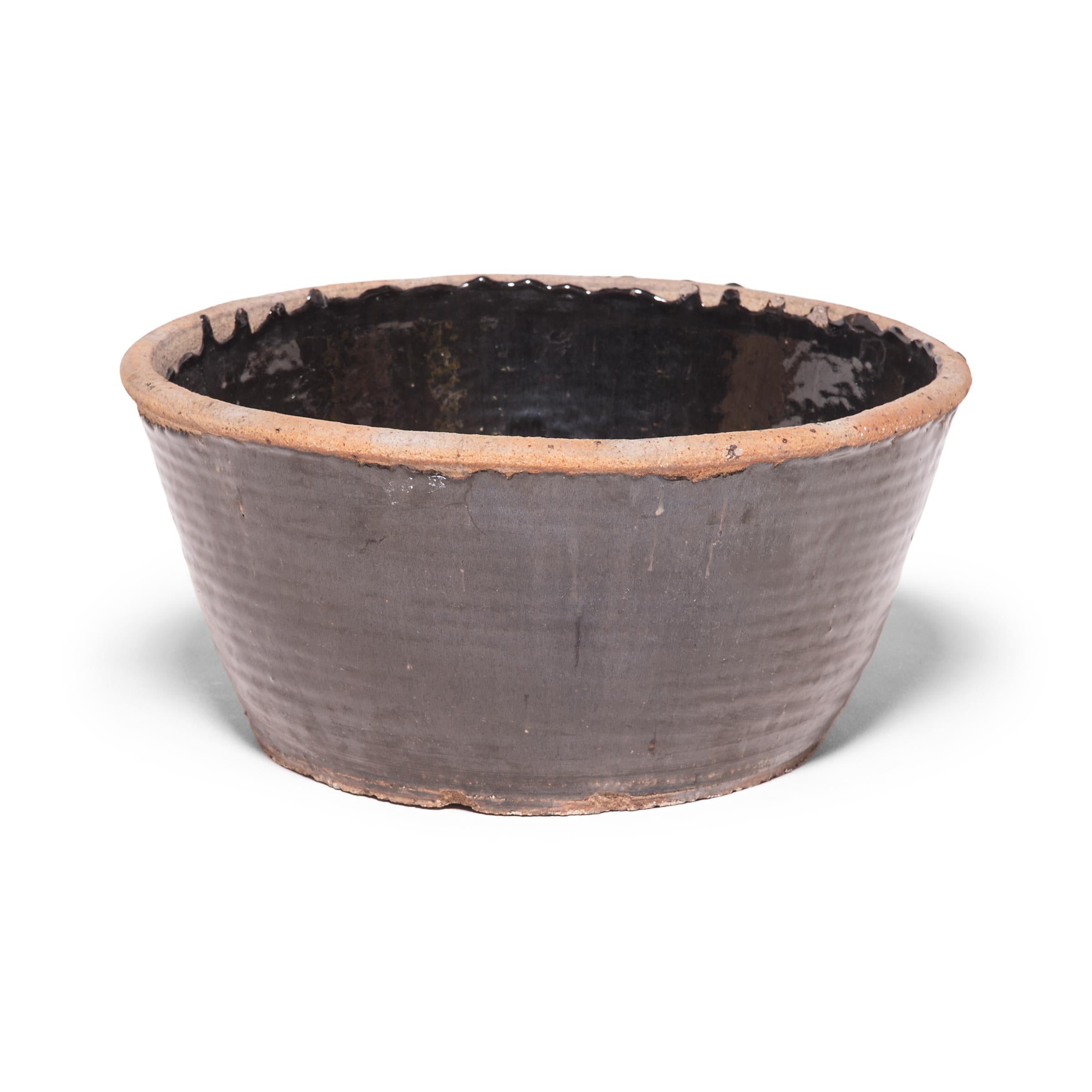 Originally used for pickling foods, this mid-20th century ceramic jar is coated inside and out with a dark glaze. Subtle ridges suggest that the bowl was fashioned by coiling flattened ropes of clay into the desired shape. Featuring an unglazed rim