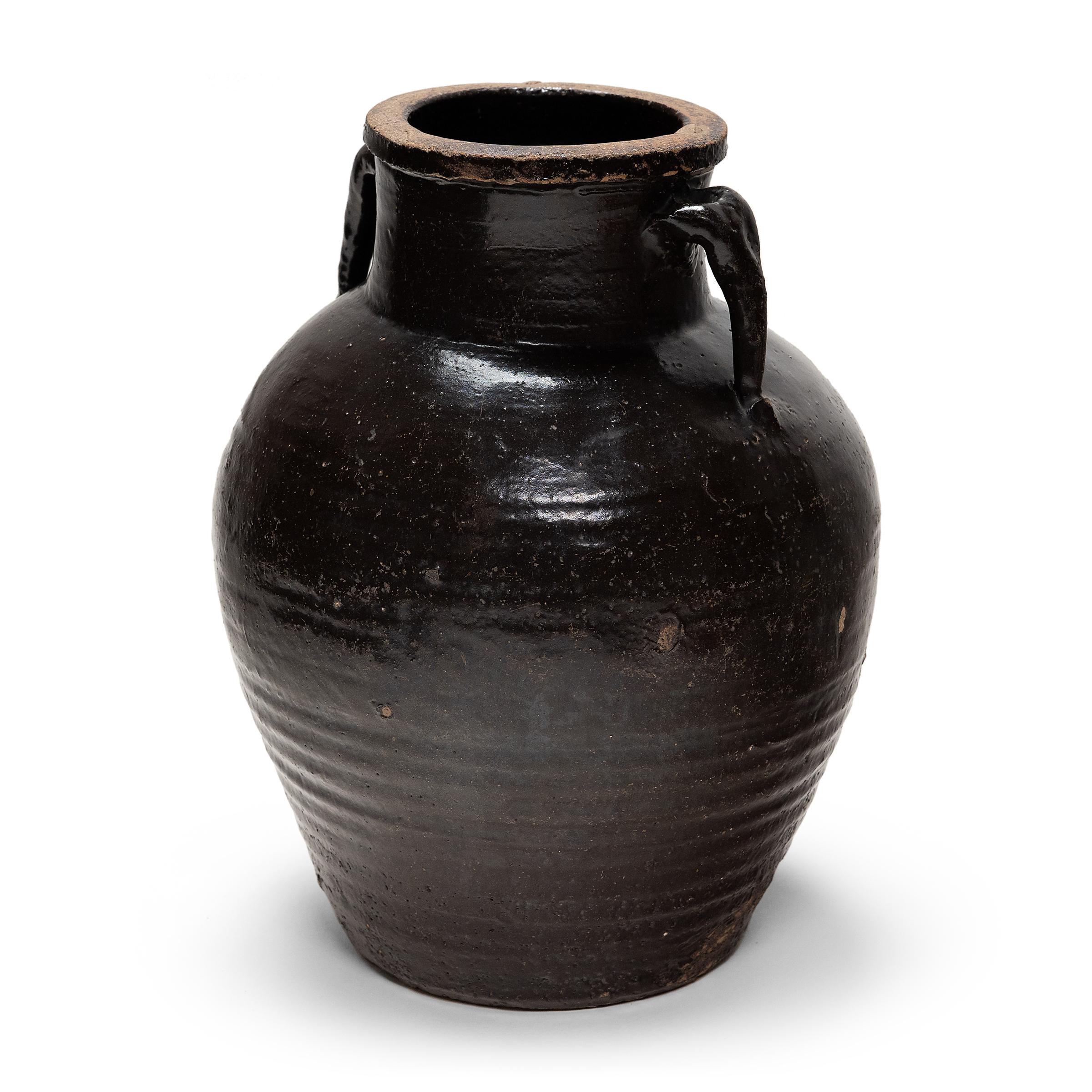 Originally used for fermenting rice or sorghum into vinegar, this earthenware vessel from the early 20th century is coated inside and out with a thick, dark glaze. The rich brown glaze sheets and pools over the jar's textured sides, lingering on