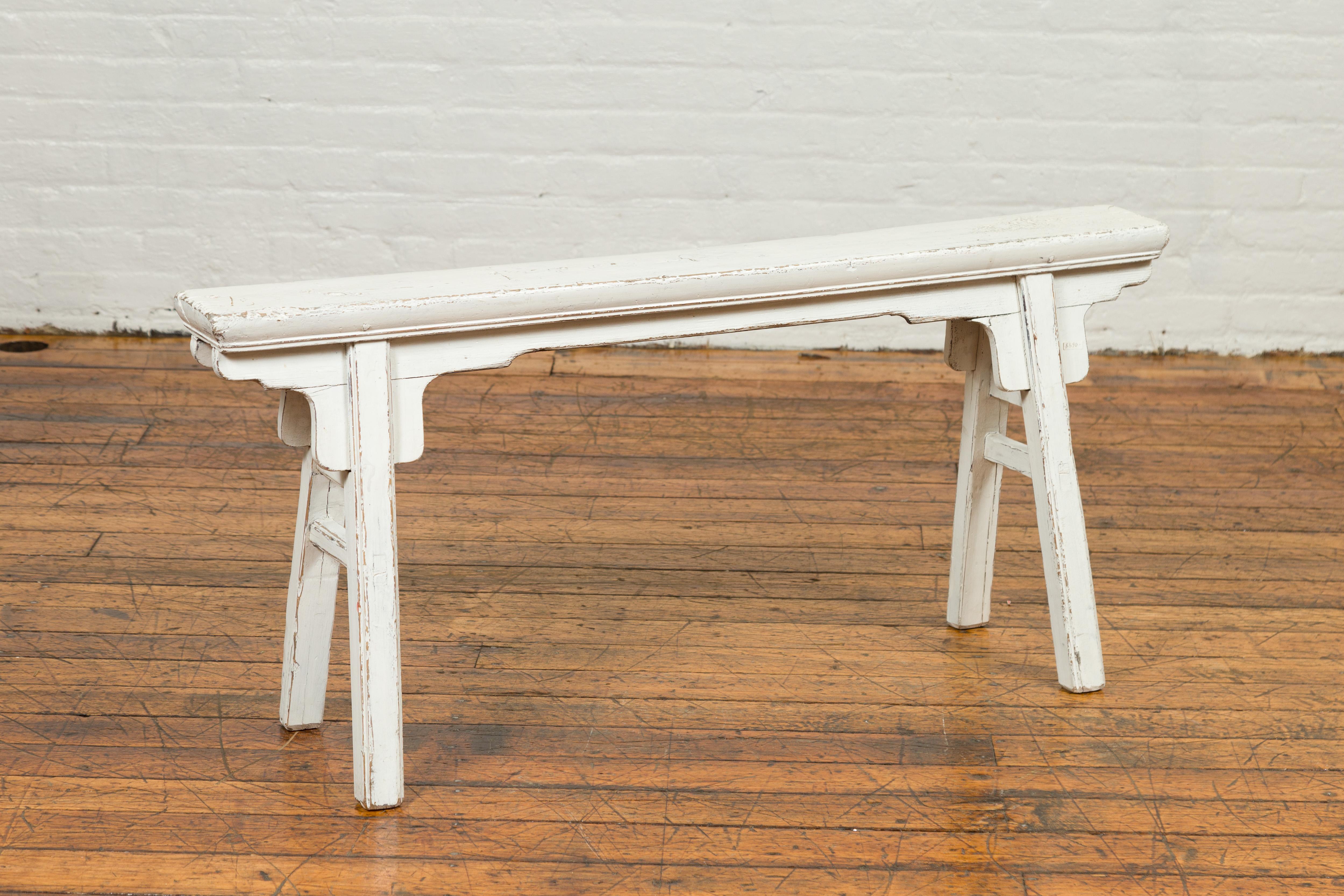 A Chinese contemporary Ming Dynasty style white painted wooden bench with A-form base and carved spandrels. Our eye is immediately drawn to the soft white finish of this Ming style bench, presenting a thin rectangular seat resting on a double A-form