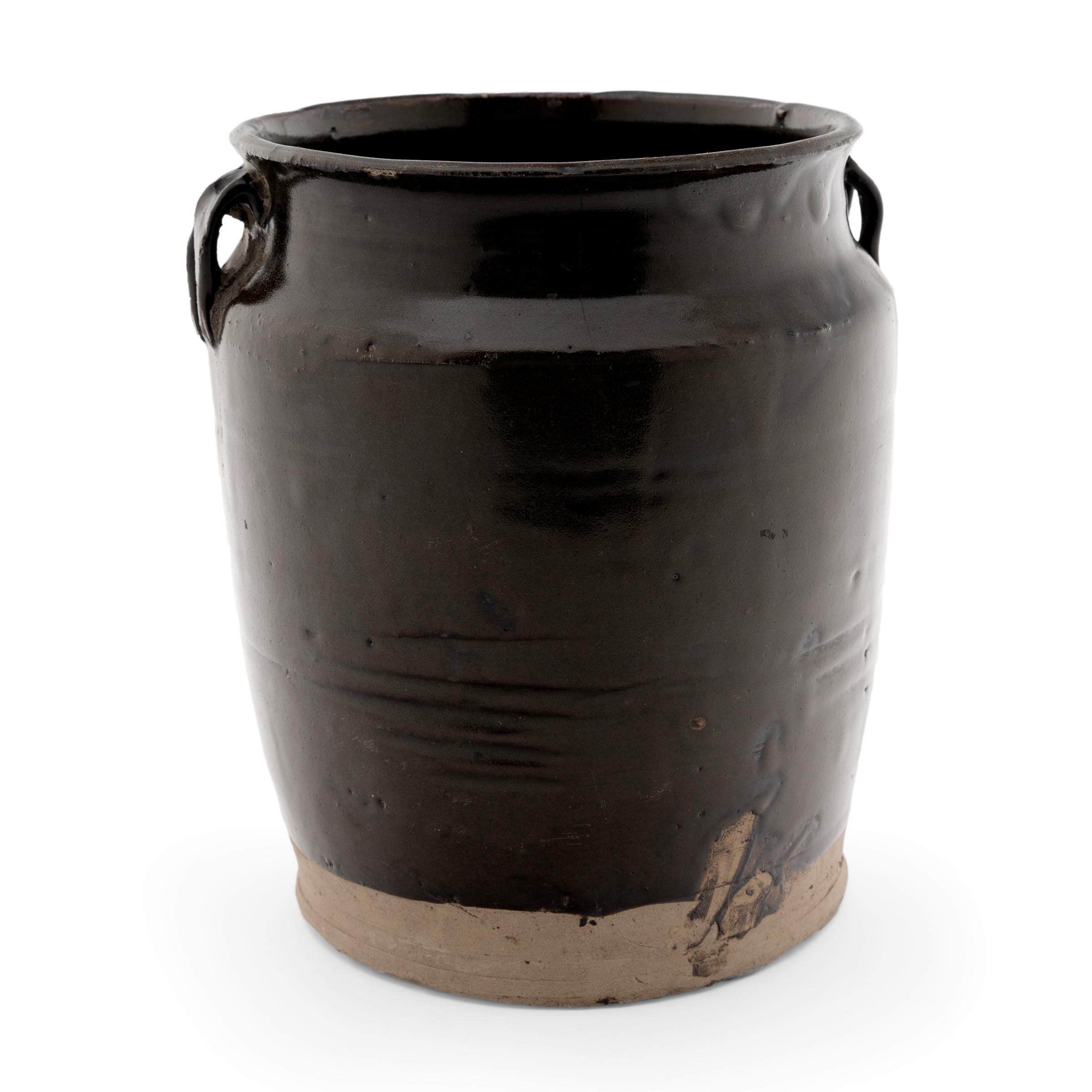 A dark brown glaze clings to the tapered form of this 19th-century jar, once used daily in a Qing-dynasty kitchen as evidenced by the glazed interior. The wide-mouth jar is shaped with gently sloping sides, rounded shoulders topped with small strap