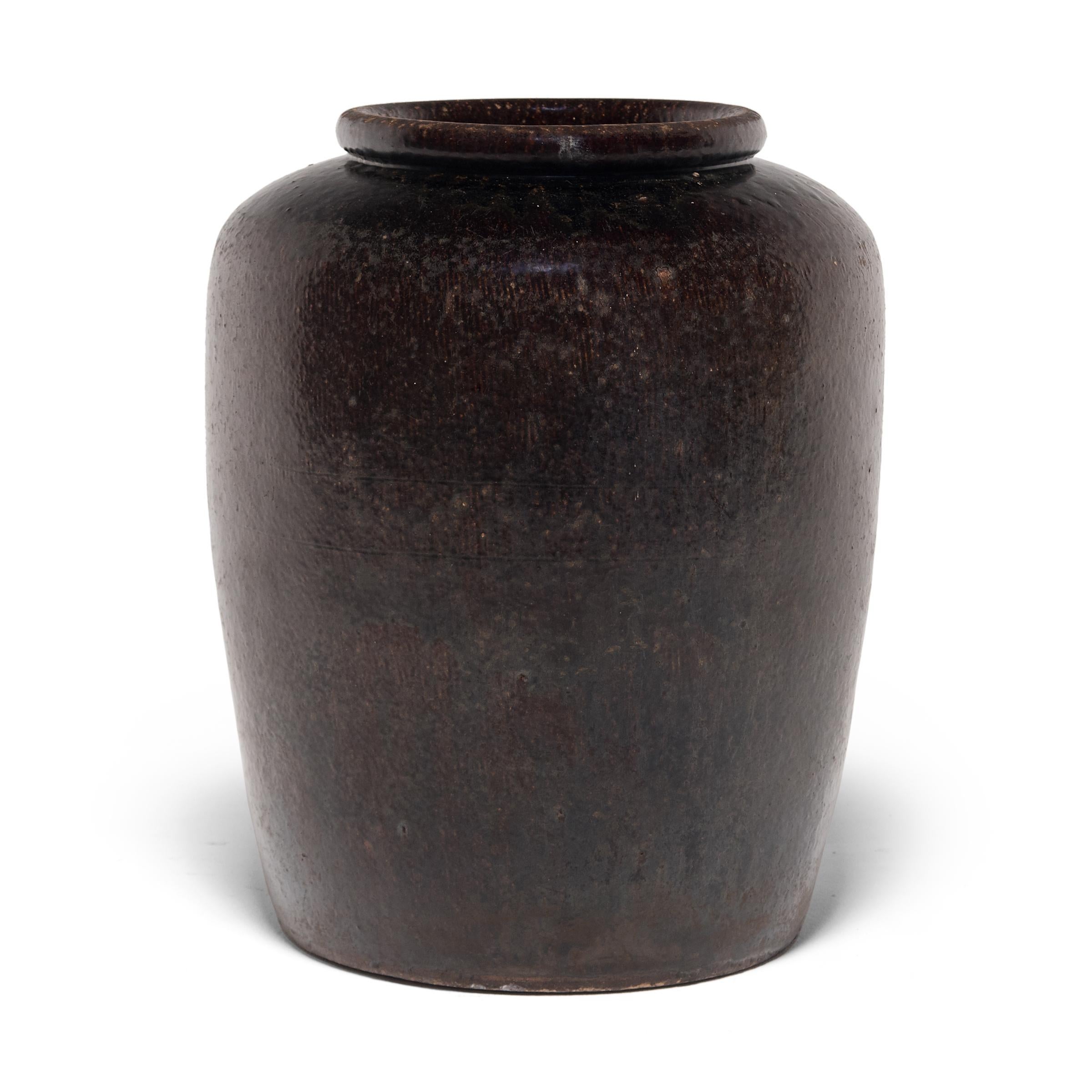 Originally used for pickling foods, this early 20th century ceramic jar is coated inside and out with a rich dark brown glaze. A subtle ridged texture patterning the jar's high shoulders manipulate the glaze to pool irregularly across the surface.