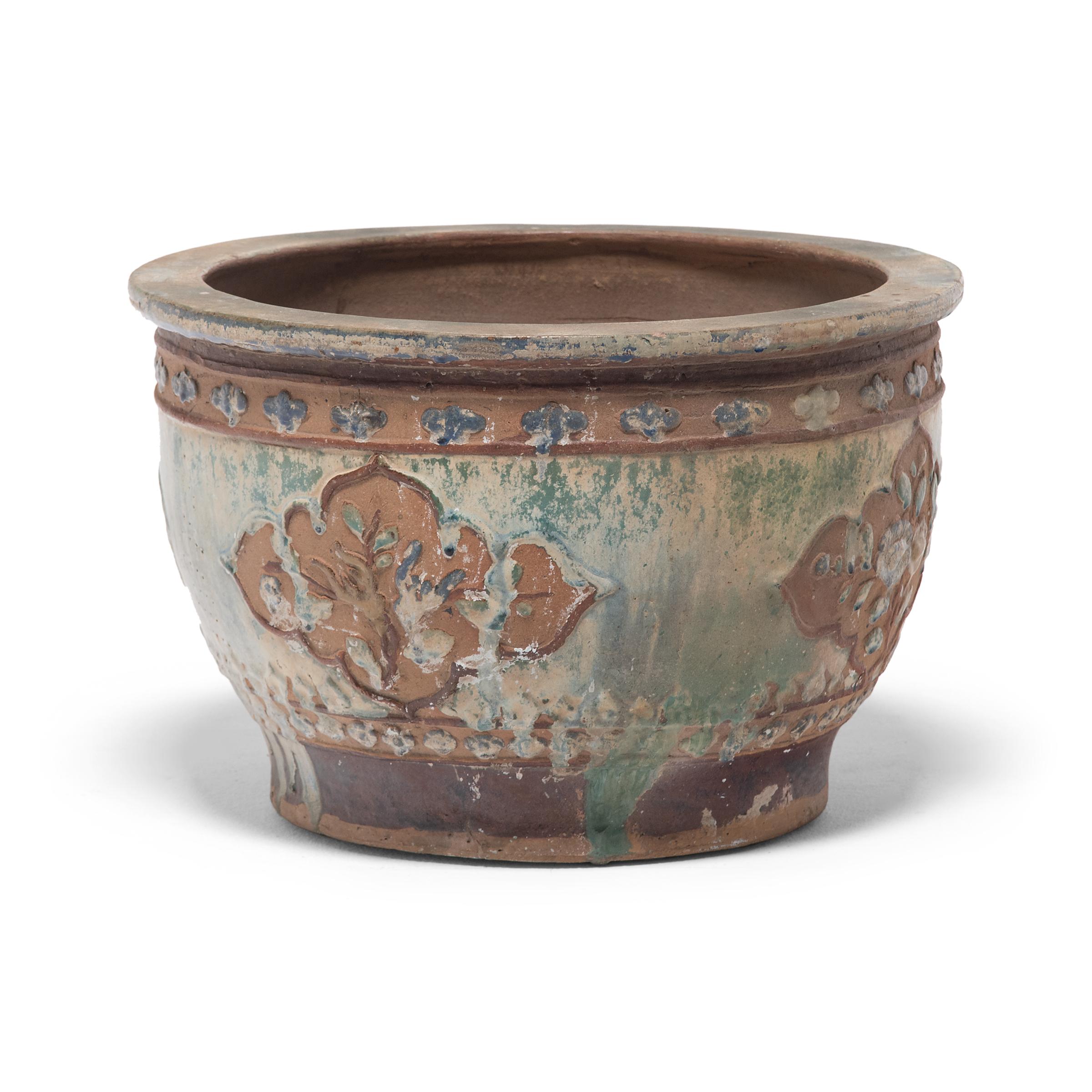 This lovely ceramic planter dates to the early 20th century and features low relief decoration and an irregular, blue-green drip glaze. The basin has a footed base, a flat lip, and rounded sides patterned with bands of simple floral motifs and lobed