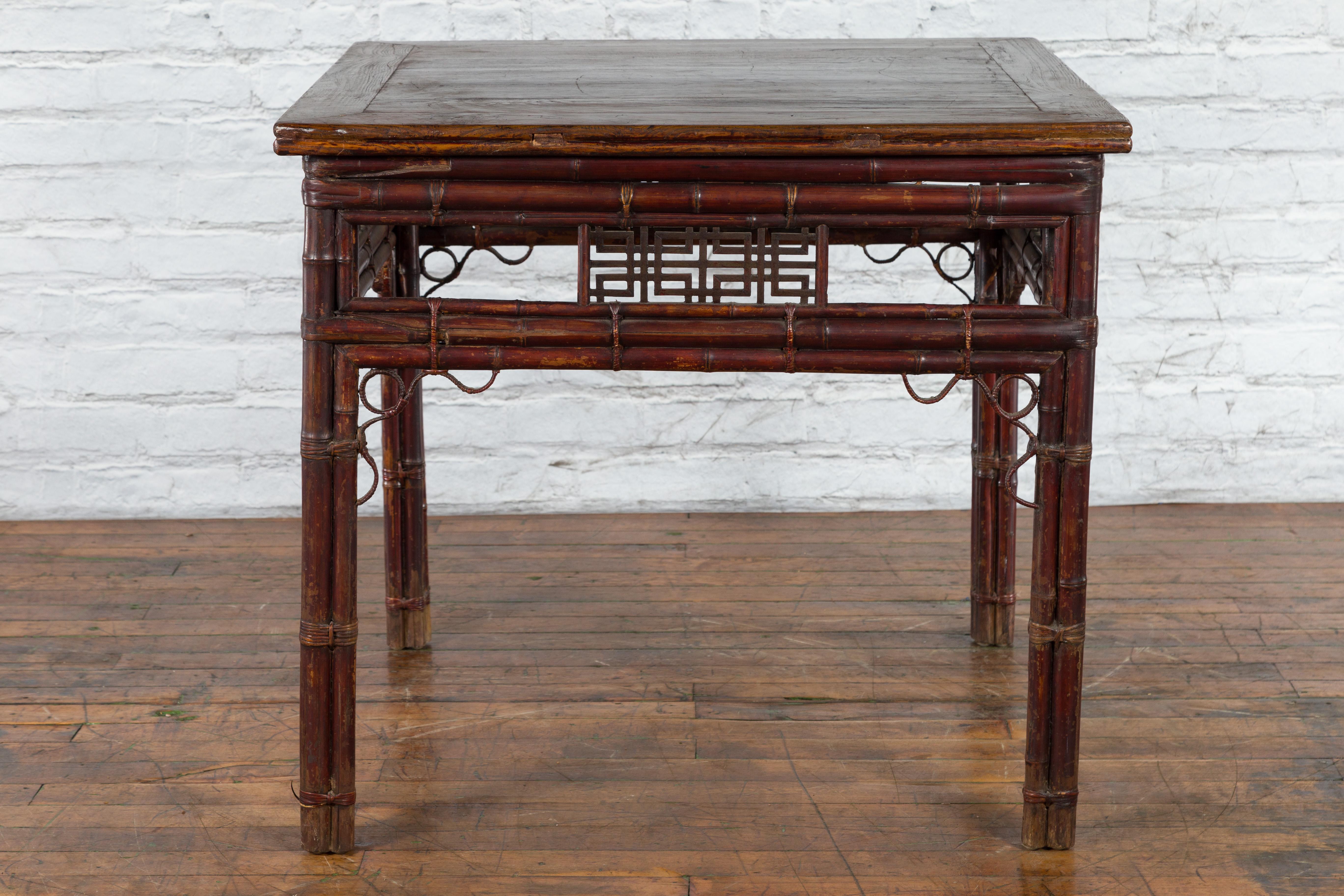 A Chinese bamboo and elm wine table from the early 20th century with fretwork motifs. Created in China during the early years of the 20th century, this wine table features a square elm top with central board, sitting above a rustic bamboo base. The
