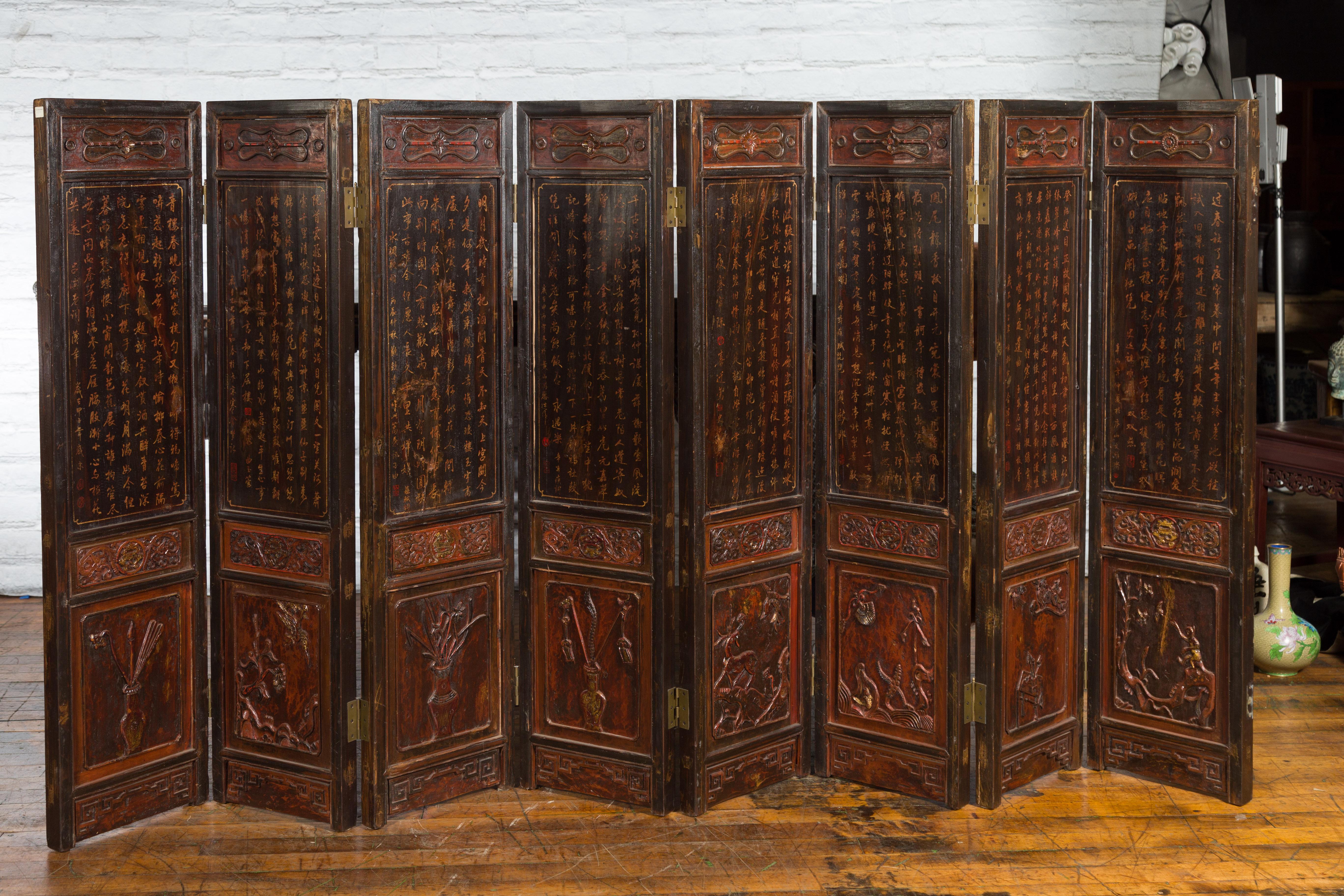 A Chinese two part eight-panel wooden screen from the early 20th century, with low-relief carvings, gilt motifs and calligraphy. Created in China during the early years of the 20th century, this screen features a brown lacquered ground accented with