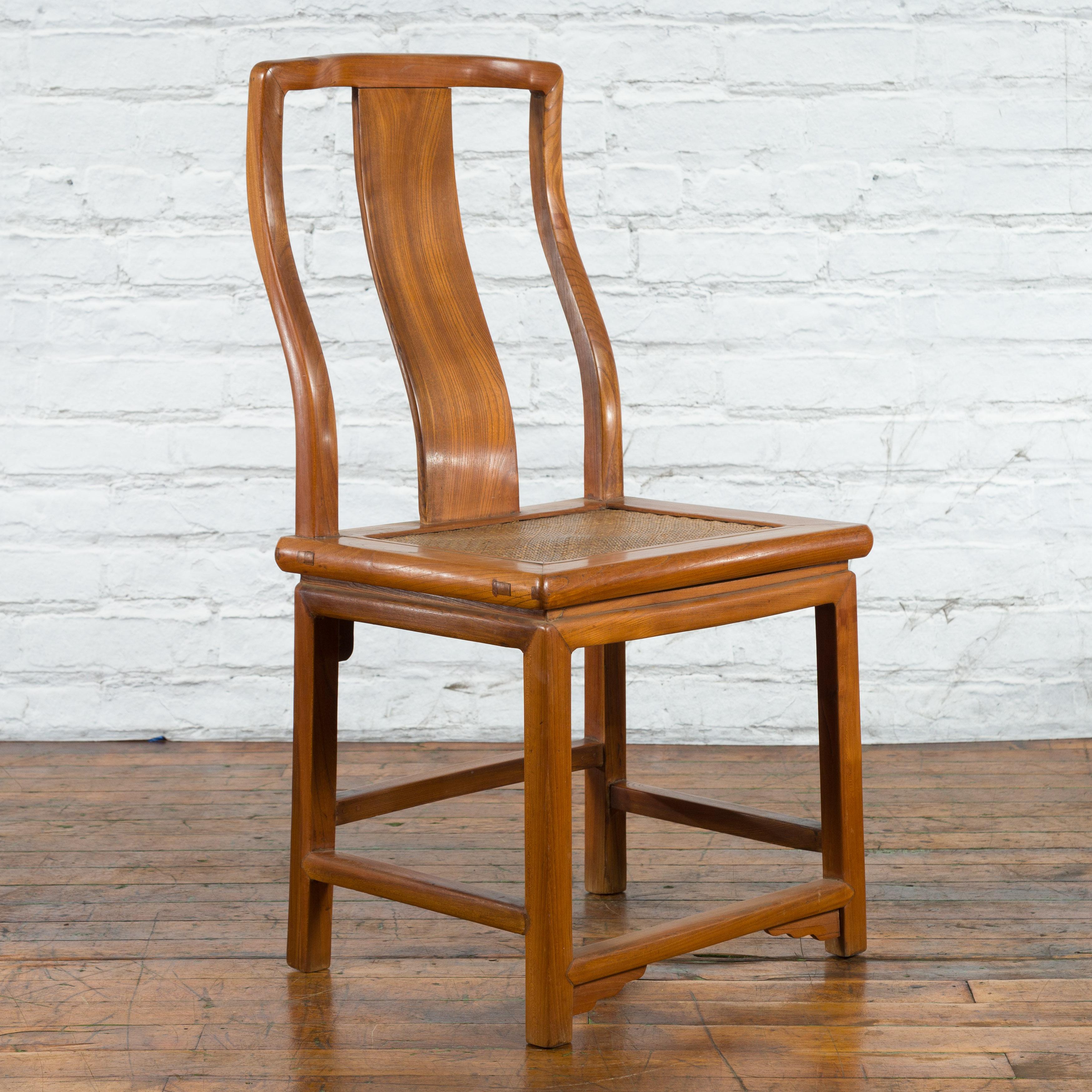 A Chinese carved elmwood side chair from the early 20th century, with woven rattan seat. Created in China during the early years of the 20th century, this elm chair features a curving back with central splat resting on a rectangular seat with rattan