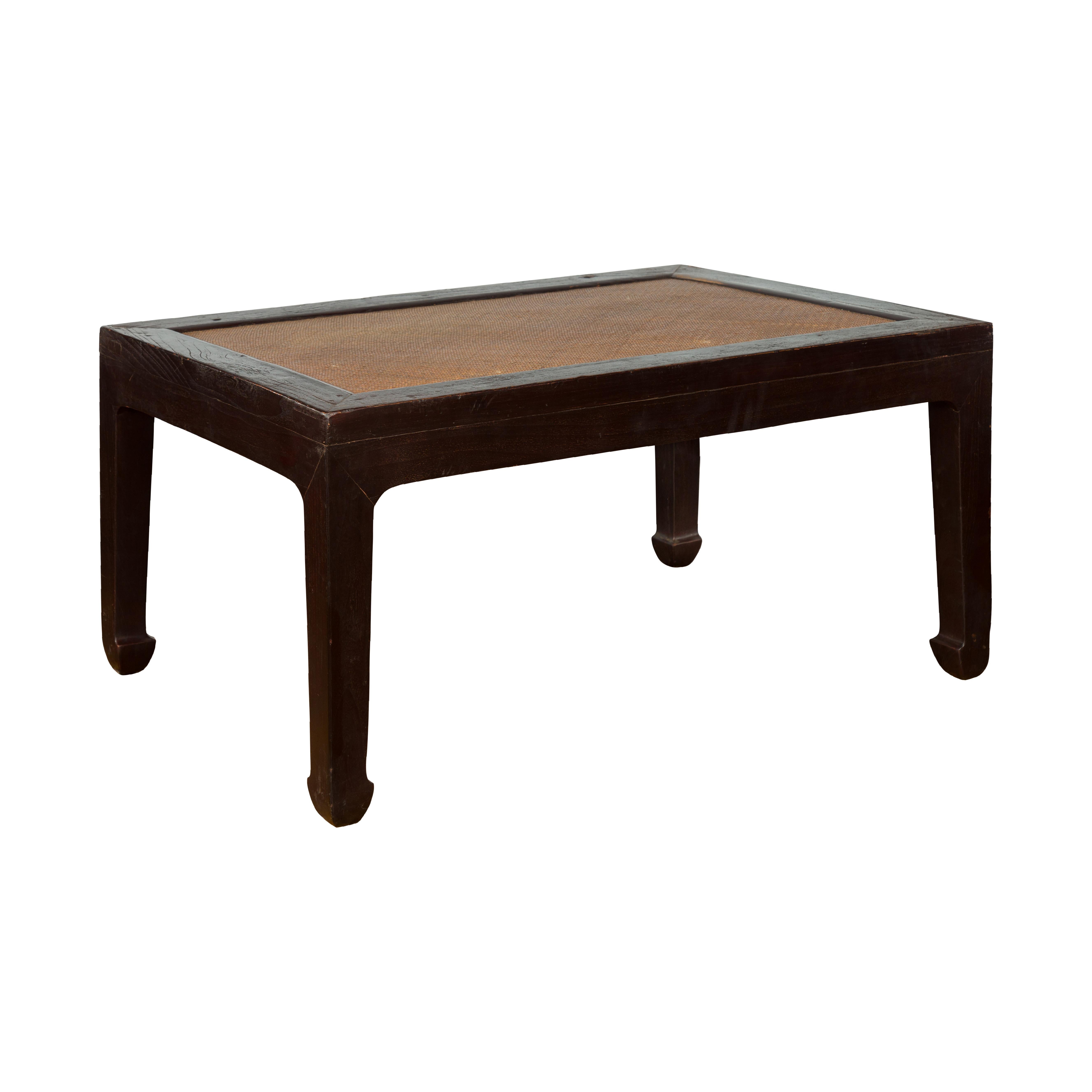 A Chinese dark brown elm wood coffee table from the early 20th century, with rattan inset top and horse hoof extremities. Created in China during the early years of the 20th century, this coffee table features a rectangular top with rattan inset