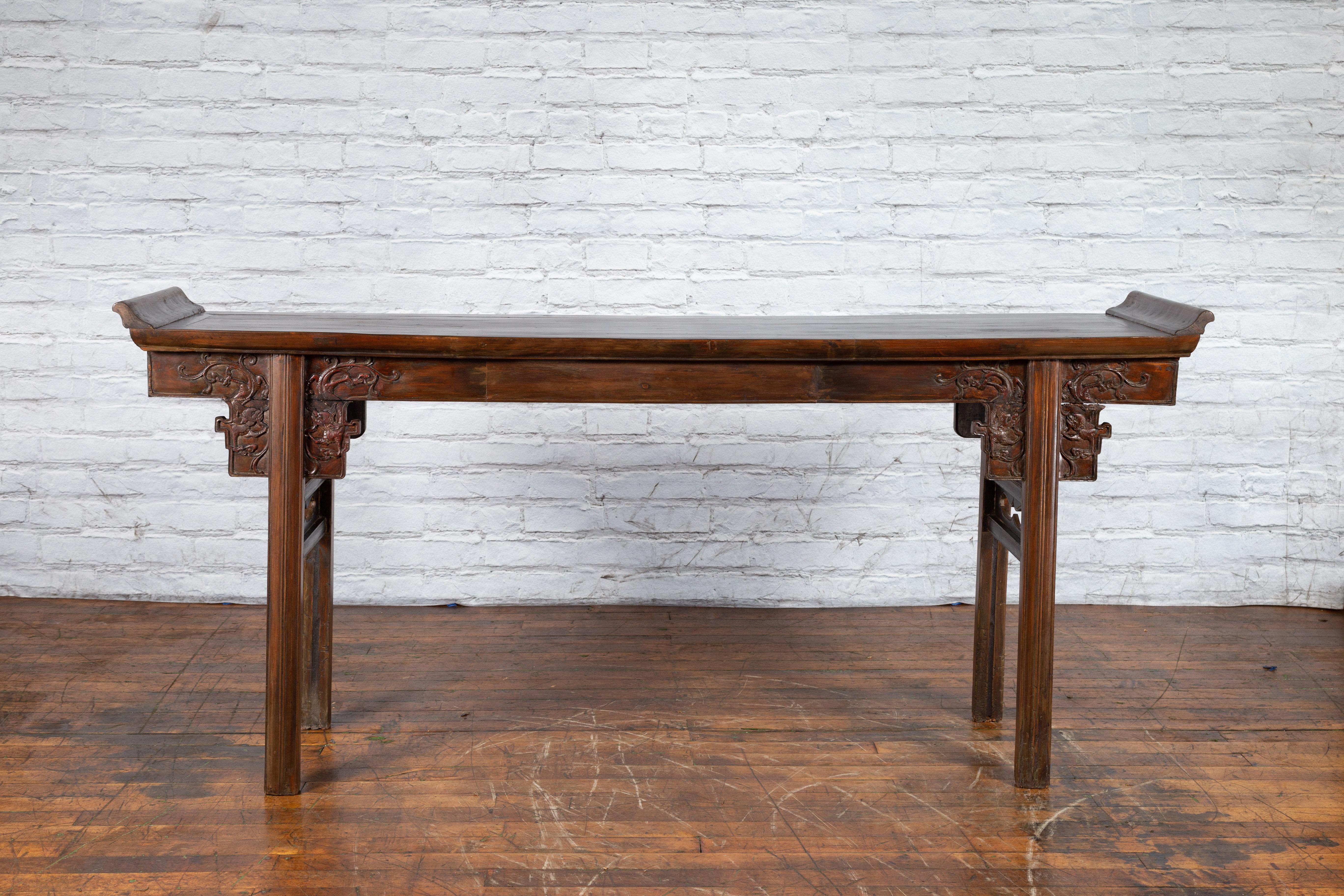 A large Chinese elmwood altar console table from the early 20th century, with carved dragons and everted flanges. Created in China during the early years of the 20th century, this altar console table features a long rectangular top flanked with