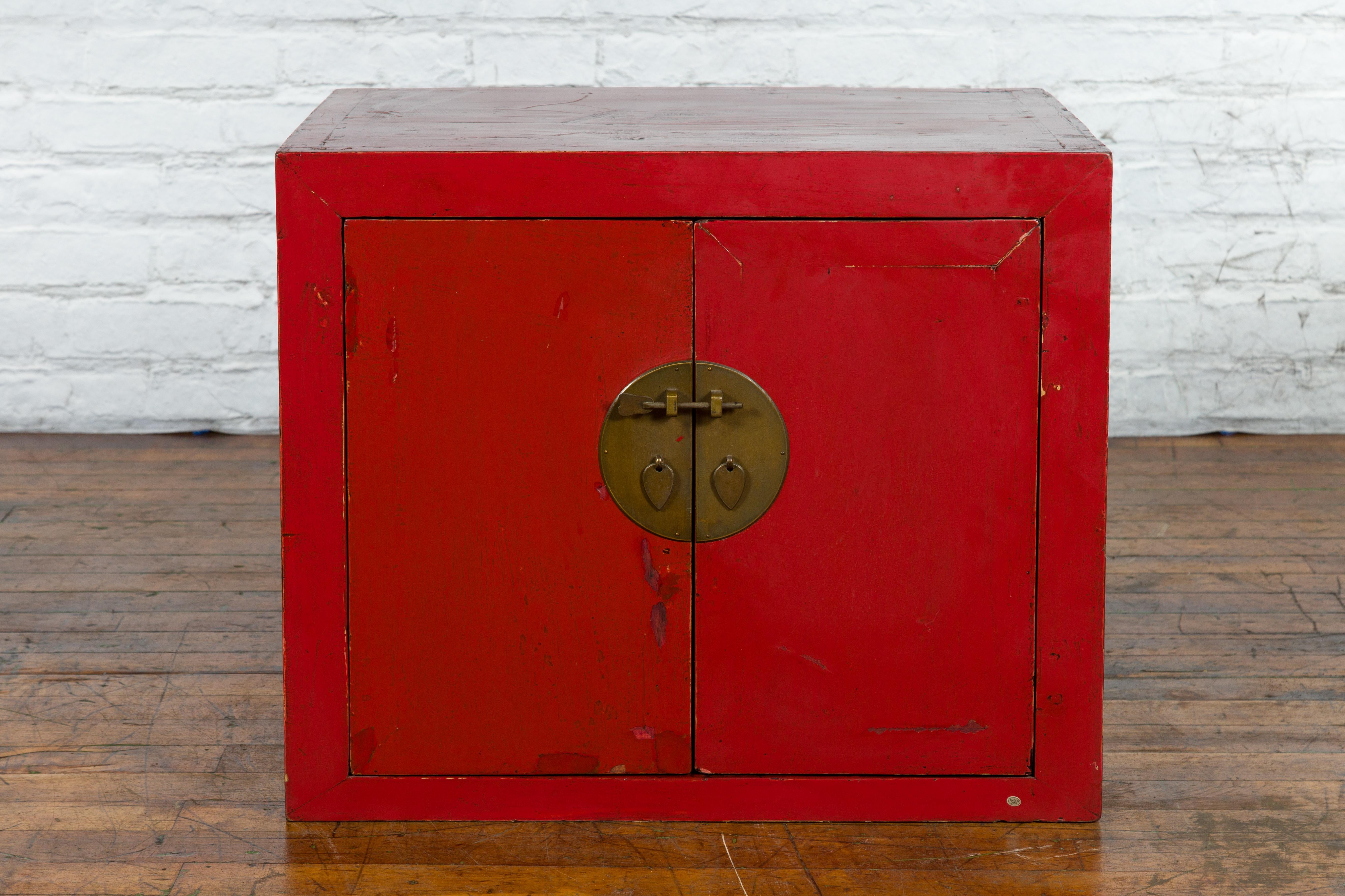 A Chinese red lacquered low cabinet from the early 20th century with original brass medallion and lock pin. Created in China during the early years of the 20th century, this low cabinet features a rectangular top with central board, sitting above