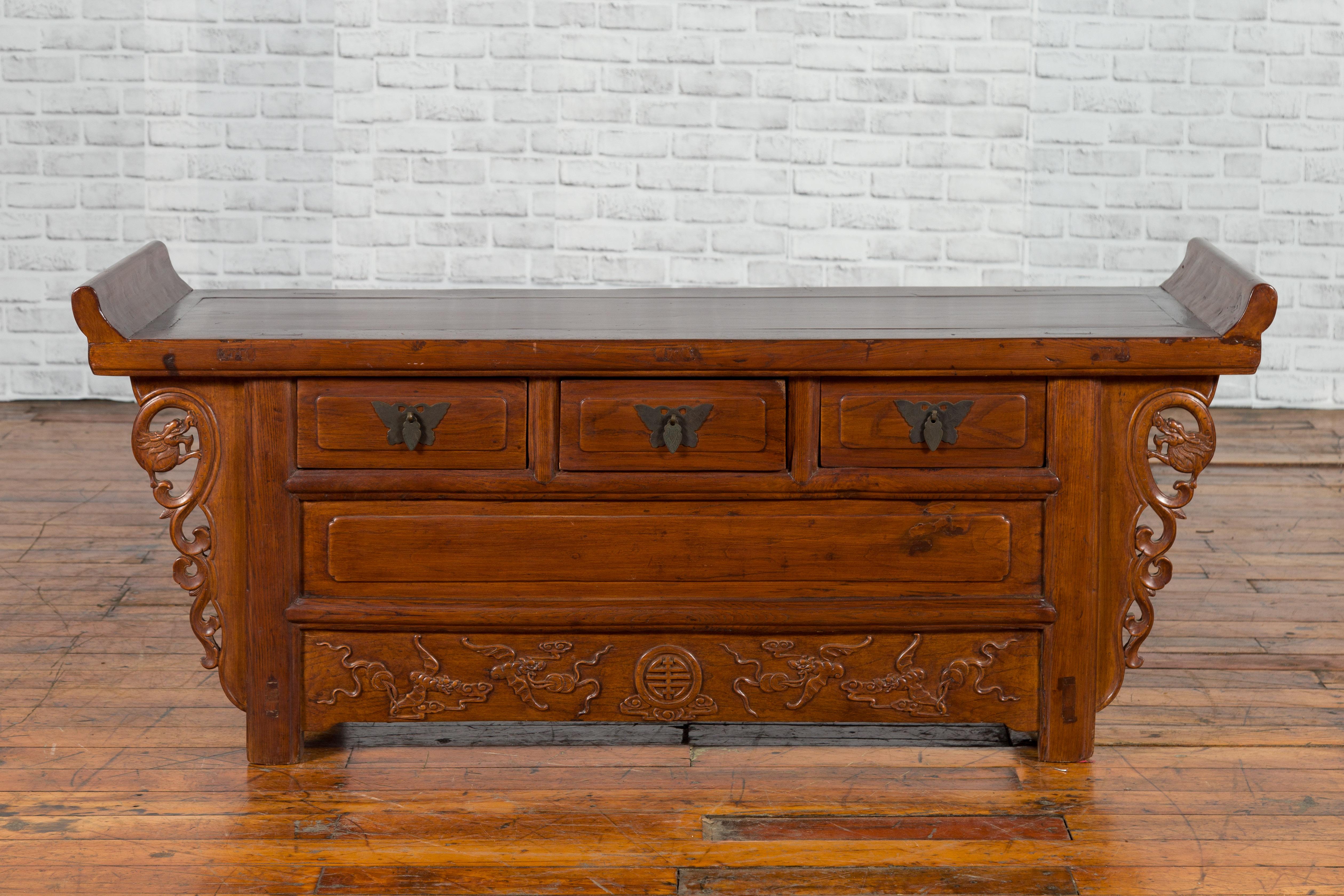 A Chinese Qing dynasty period elm kang cabinet with everted flanges, three drawers, carved spandrels and dragon motifs. Created in China during the Qing dynasty, this Chinese kang cabinet features a rectangular top with central board, flanked with