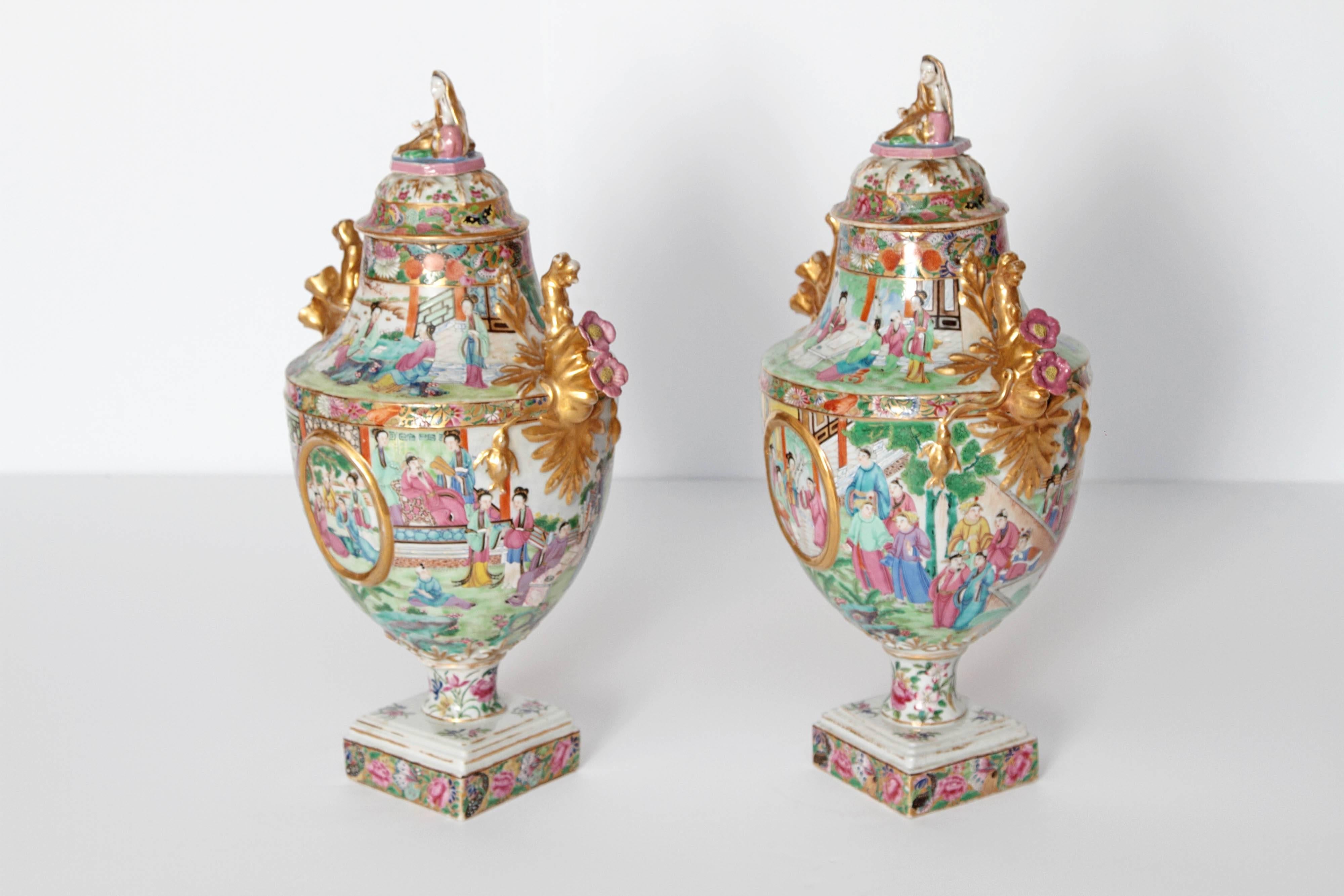 A pair of Chinese covered urns with famille rose coloration with all-over pattern of people and floral decoration, elaborate gold handles with pink and yellow flowers, additional gold / gilt ovals / medallions on both fronts and backs, lids are