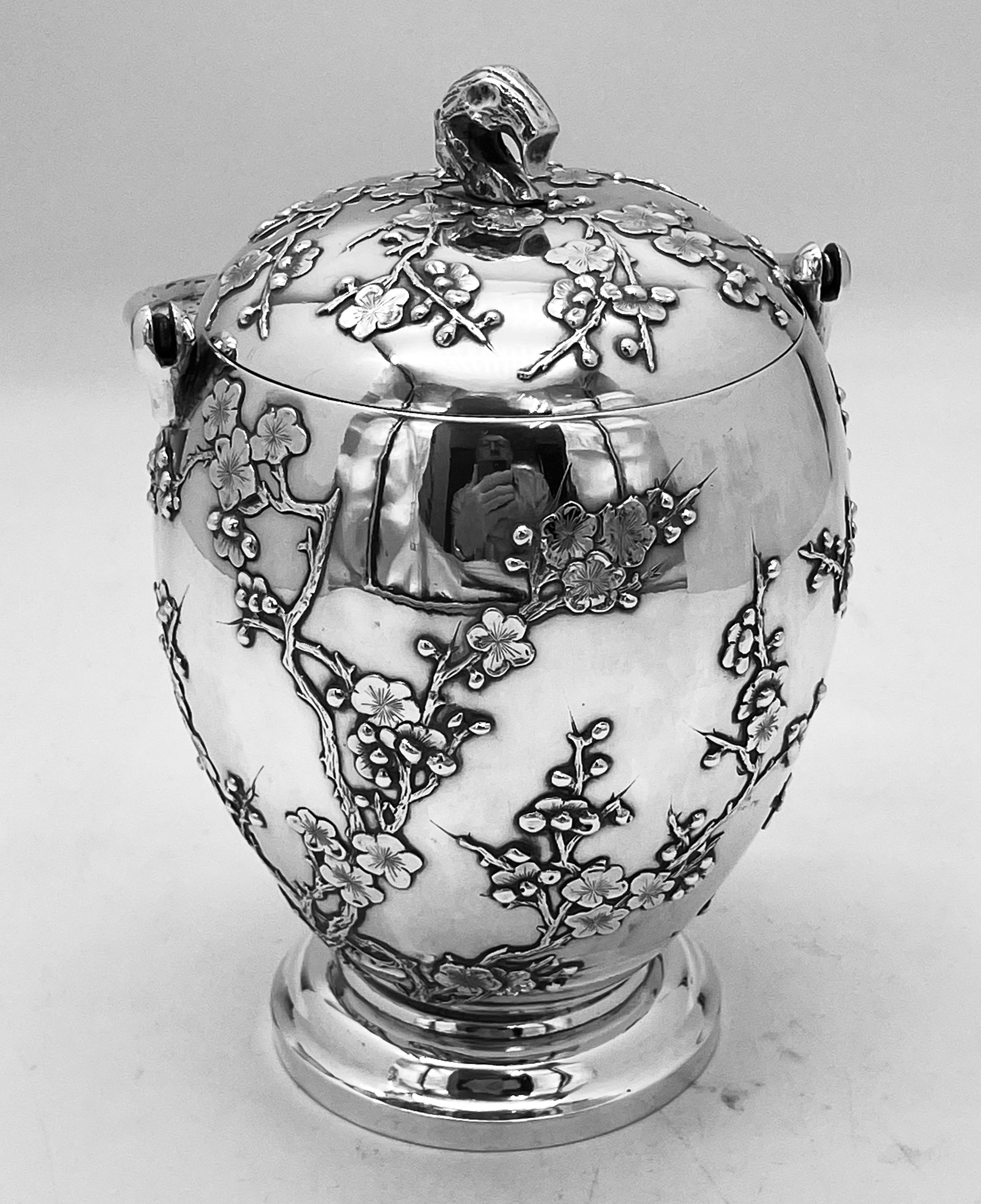 A Chinese Export Silver Biscuit Box, round with a collet foot and profusely decorated with applied prunus. against a smooth plain background. The box has a pull-off lid with a twig finial, and a plain swing handle.
Whilst originally made to hold