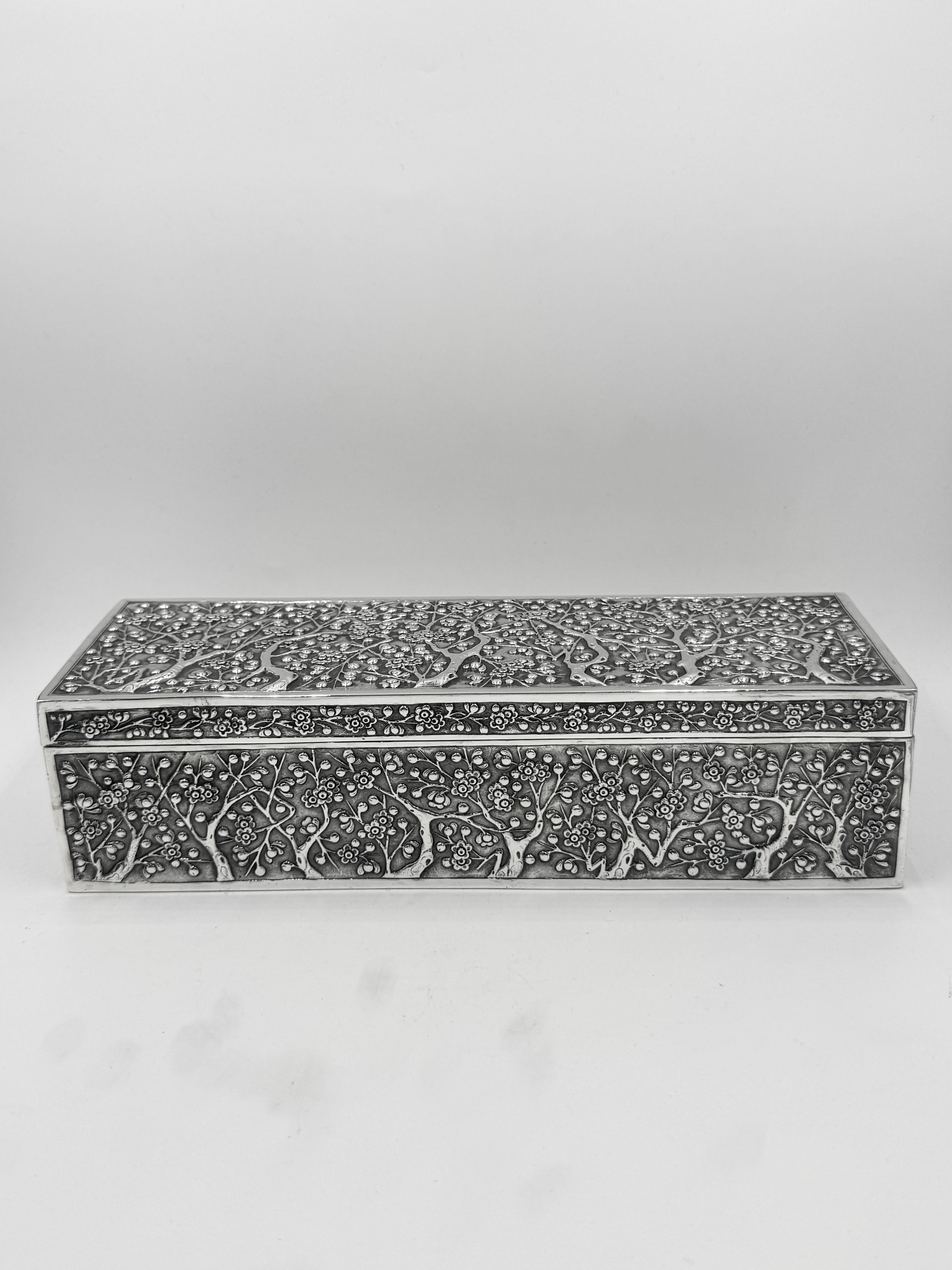 Scatola da tavolo in argento da esportazione cinese del 1890 circa di forma rettangolare allungata, con rami di pruno e fiori in rilievo su uno sfondo opaco. 
La scatola è contrassegnata dalla sigla WH, che sta per Wang Hing. Essendo la più grande
