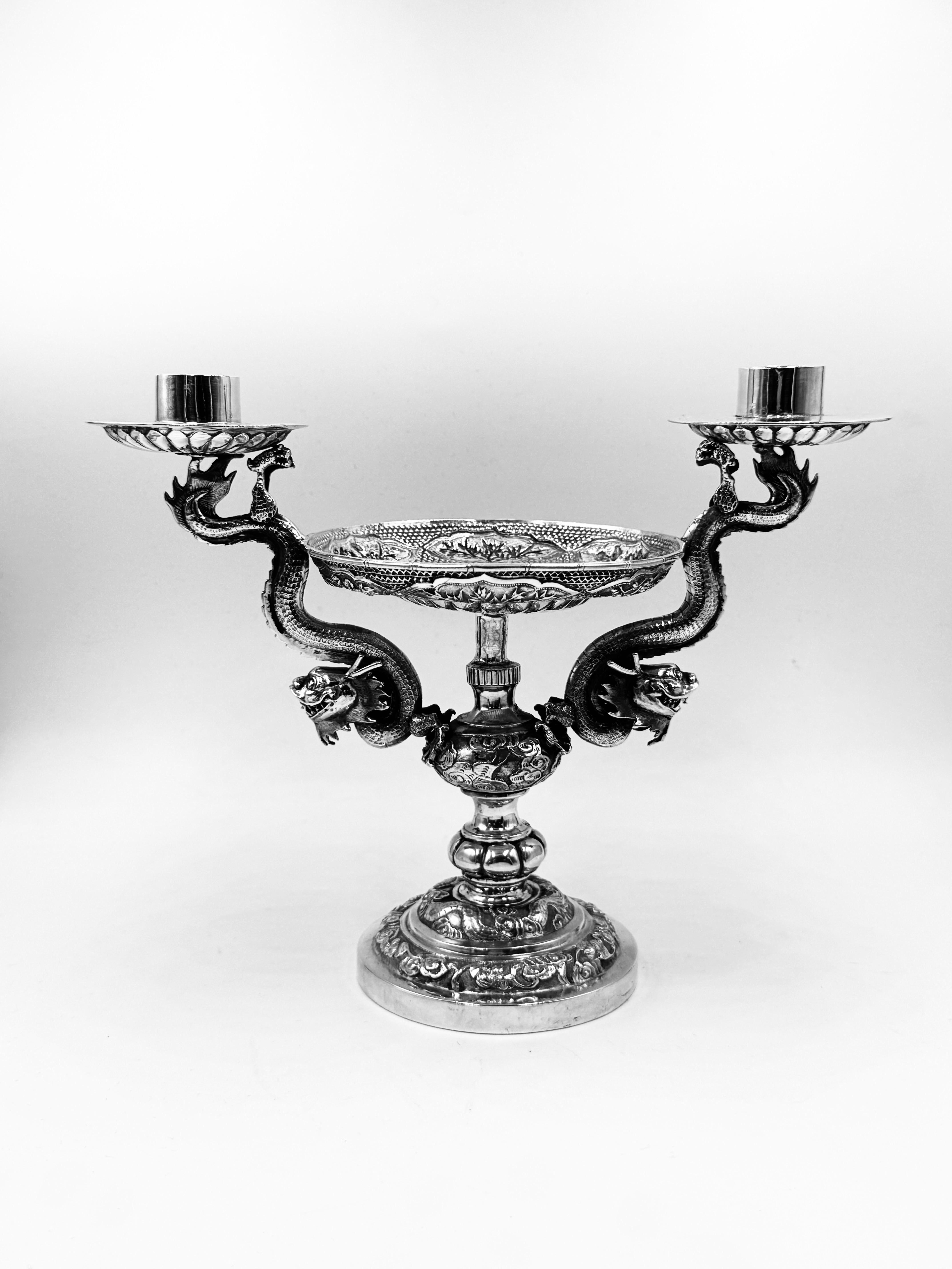 Centro de mesa chino de hacia 1895, en forma de candelabro de 2 luces con un plato central sostenido por dos dragones voladores. 
La parte inferior de los portavelas y el plato están estriados, y la base y la columna están repujadas y cinceladas con