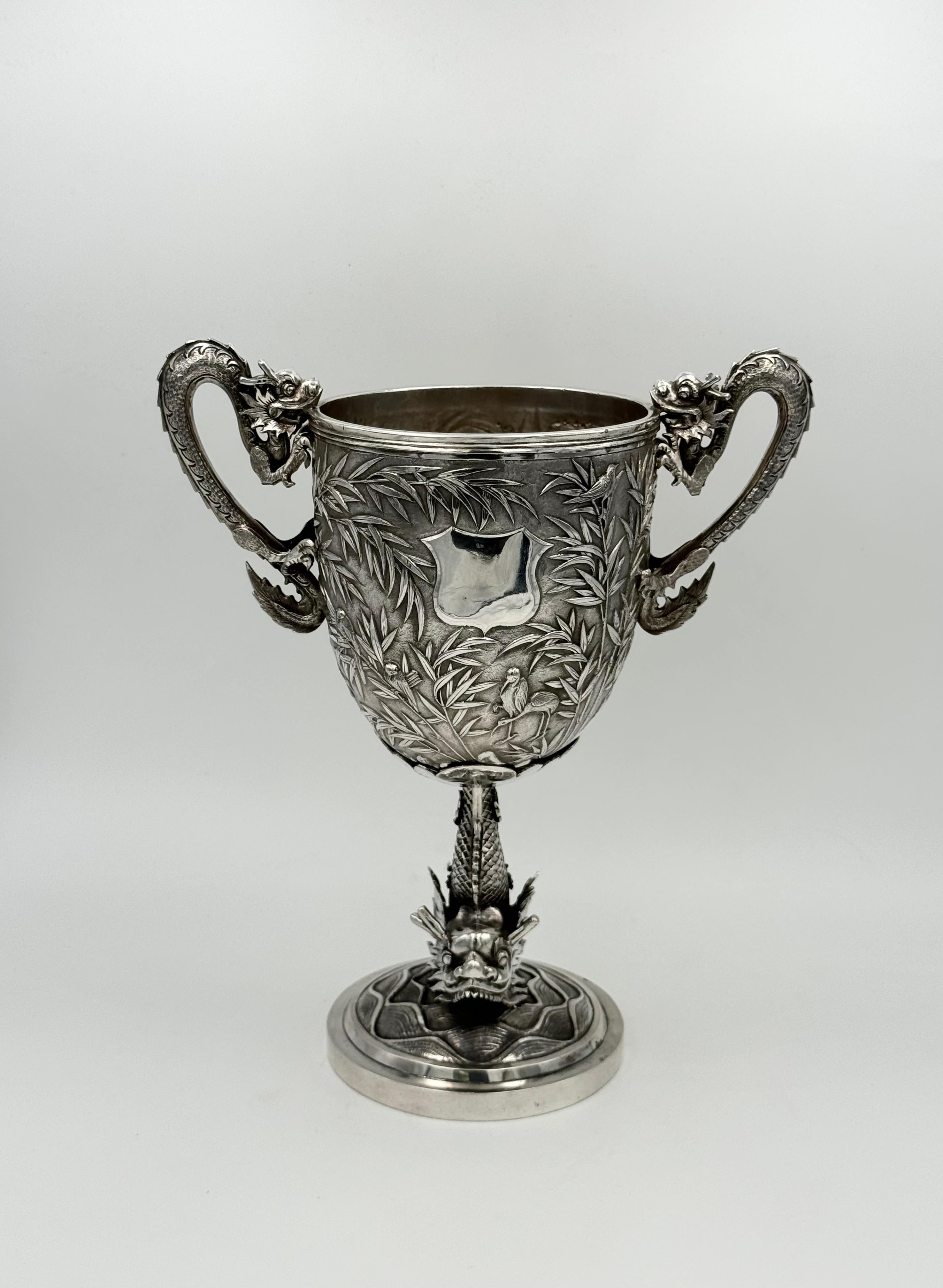 A Chinese Export Silver Cup with dragon handles, and with birds and bamboo chased and embossed around the body. The cup is supported by a further dragon atop a base of waves. With a mark for Wang Hing and dating from around 1890.
