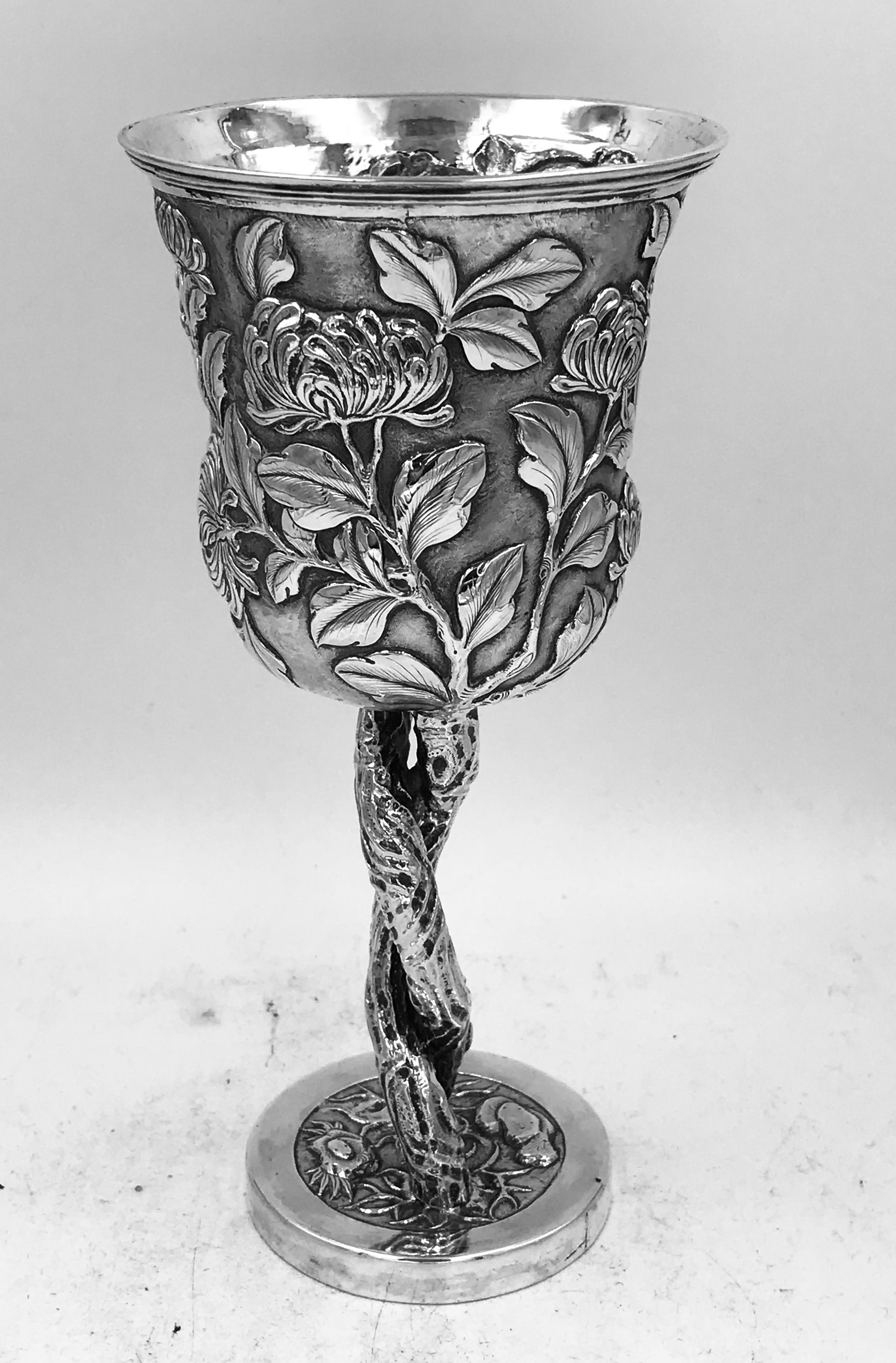 A Chinese silver goblet profusely decorated with chased, richly defined chrysanthemum against a matte background and is supported by two intertwined branches forming the stem. It was made by Yuan Xin circa 1890. 
The goblet is 188mm high and weighs