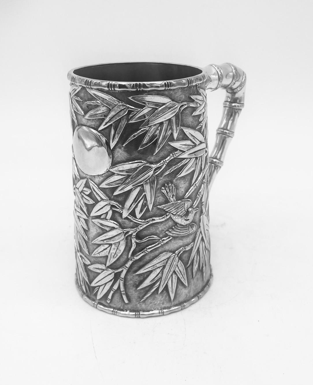 A Chinese Export silver mug with a bamboo handle, and decorated with a bird and moth amongst profuse bamboo decoration on a matte background. The mug is of double-skinned form and the interior sleeve has traces of old gilding. 
The total weight is