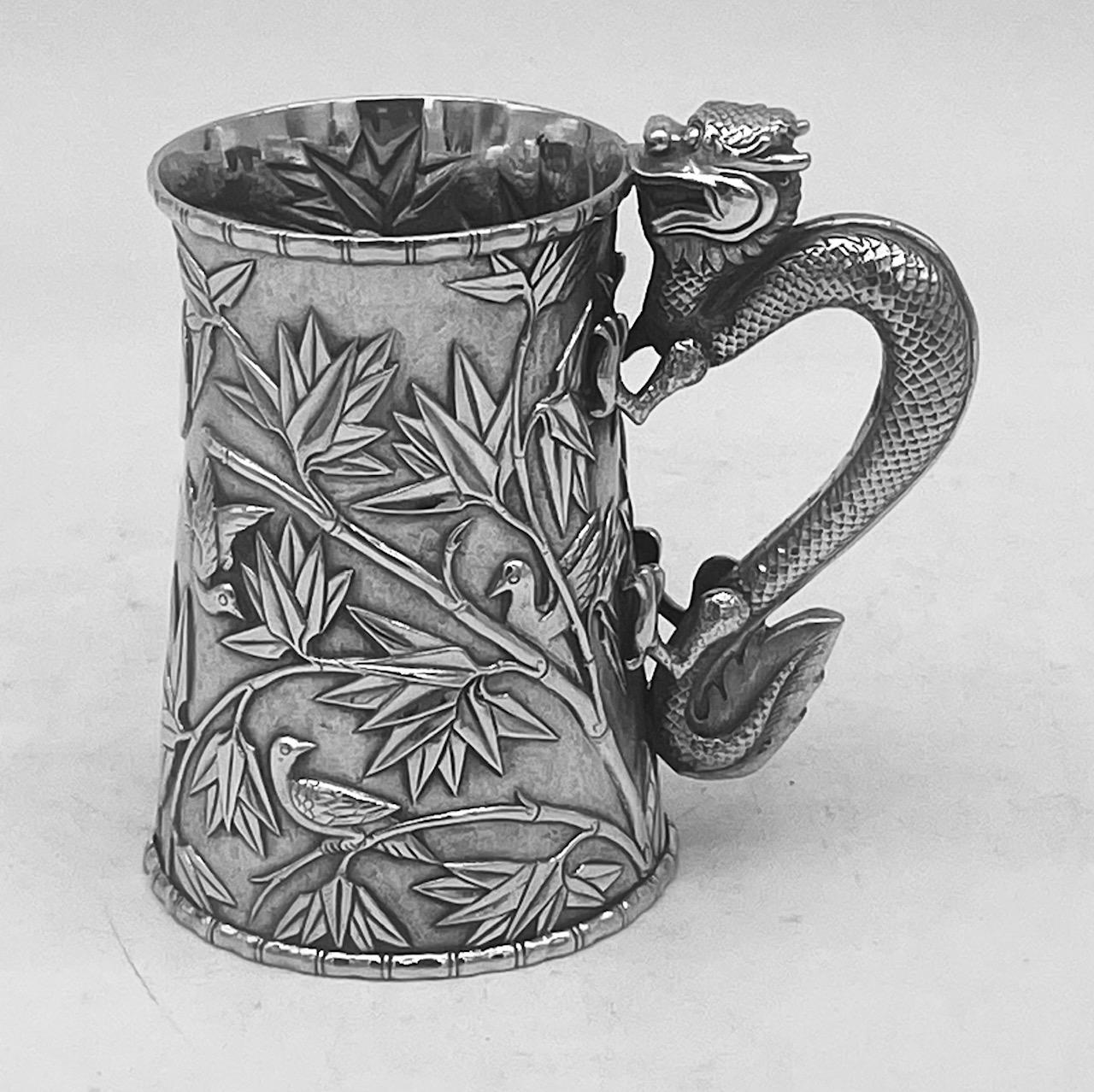 A Chinese Export silver mug with dragon handle, decorated with five birds among bamboo against a matte background. There is a round vacant cartouche to the front and the inside has traces of old gilding. 
The base of the mug is marked LEO, which