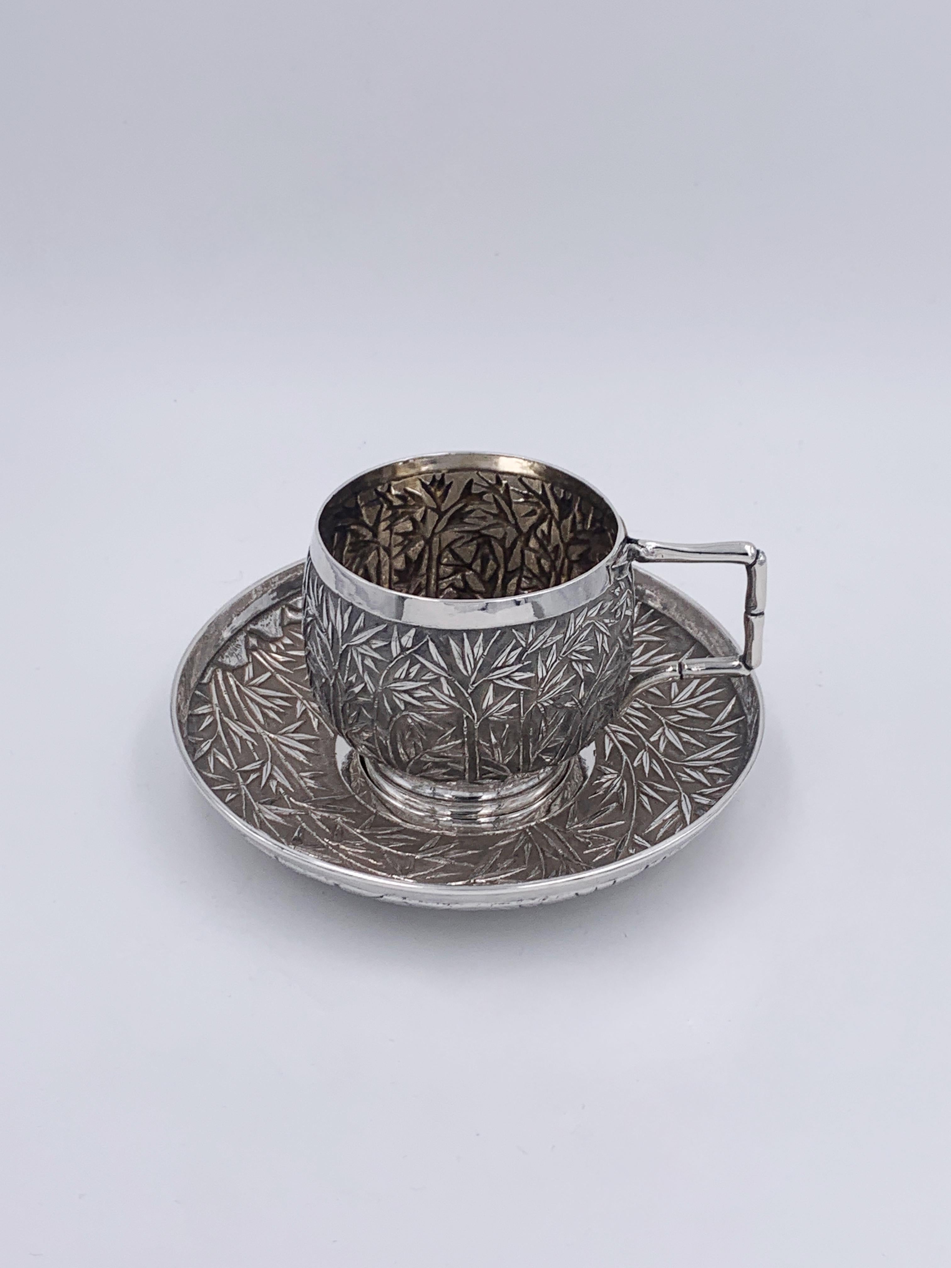 A Chinese Export Silver Tea Cup and Saucer embossed and chased with bamboo against a matte background. The cup has a bamboo-form handle and sits on a plain collet foot, and the saucer has the same decoration.The retailer marked as WH for 'Wang Hing'