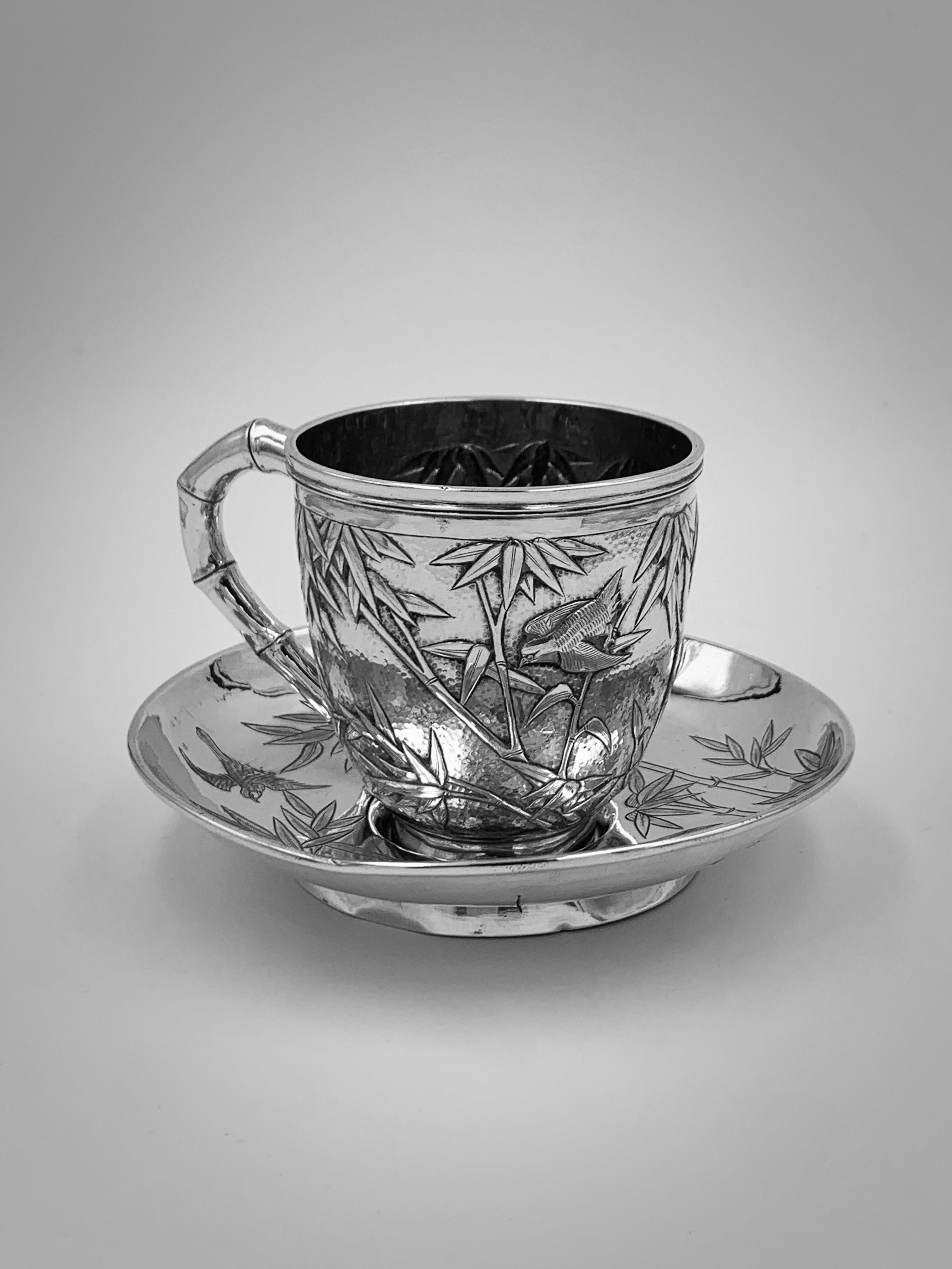 A Chinese Export Silver Tea Cup and Saucer made in Hong Kong around 1900. The cup embossed and chased with birds and bamboo against a matte background, with a single vacant shield-shape cartouche and a bamboo-form handle. The saucer is engraved with