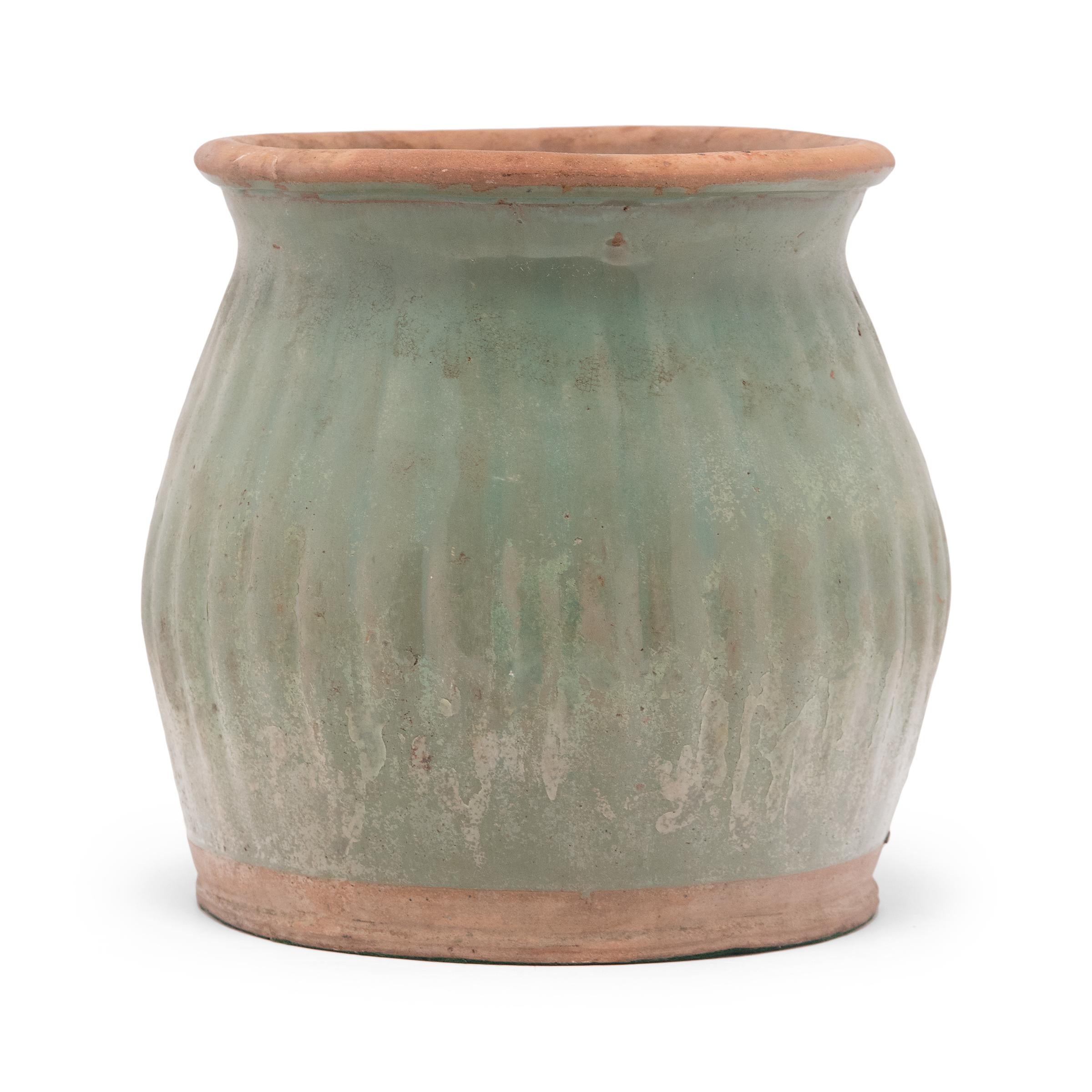 Dated to the early 20th century, this glazed ceramic jar features a flared lip and bulbous sides textured by a pattern of ridges. A celadon-green glaze coats the swelling sides, beautifully aged with minor crazing and pitted wear. The top opening is