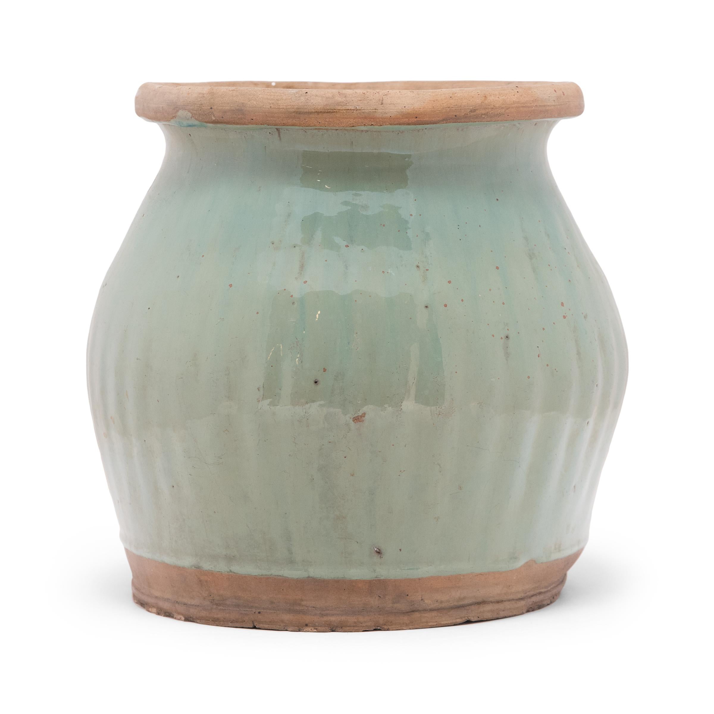Dated to the mid-20th century, this glazed ceramic jar features a flared lip and bulbous sides textured by a pattern of ridges. A celadon-green glaze evenly coats the swelling sides, dotted with pitted wear and subtle streaks of blue. The top