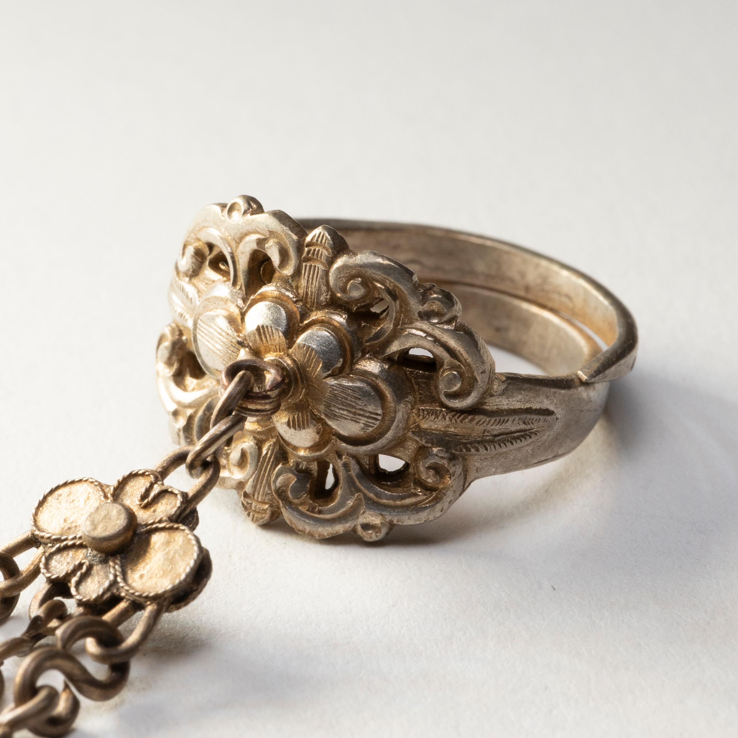 Datée de la fin du XIXe siècle, cette bague à breloques en argent était censée protéger son porteur de la malchance et des esprits malveillants. L'anneau est décoré en relief d'une fleur stylisée entourée de feuilles et de rinceaux. Trois chaînes