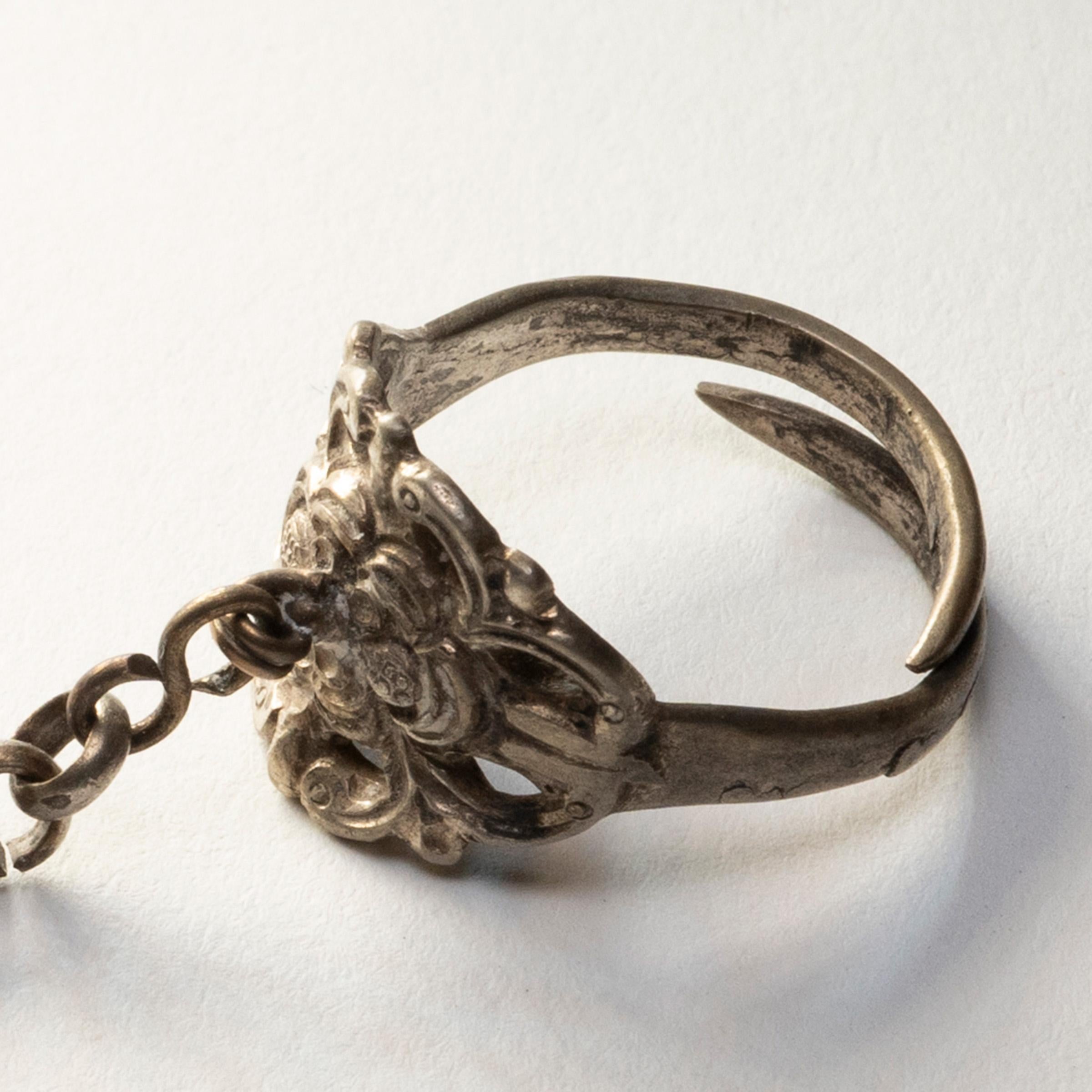 Dated to the late 19th century, this silver charm ring was believed to protect the wearer from bad luck and malevolent spirits. The ring is decorated in relief with a flower blossom surrounded by leaves and curling tendrils. A chain hangs from the