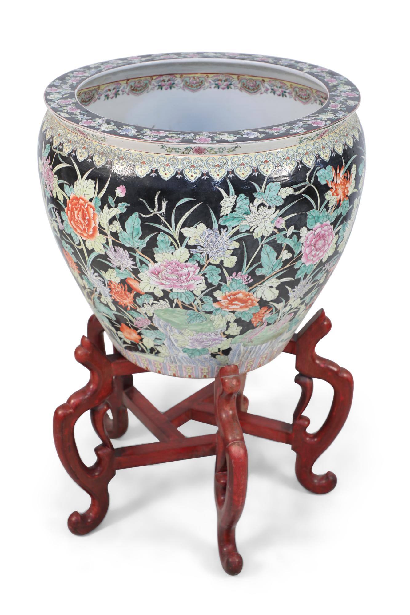 Chinese porcelain planter painted with florals and greenery over a black ground on the exterior and fish amidst seaweed along the interior. The pot sits atop a 5-footed wooden stand connected by stretchers.