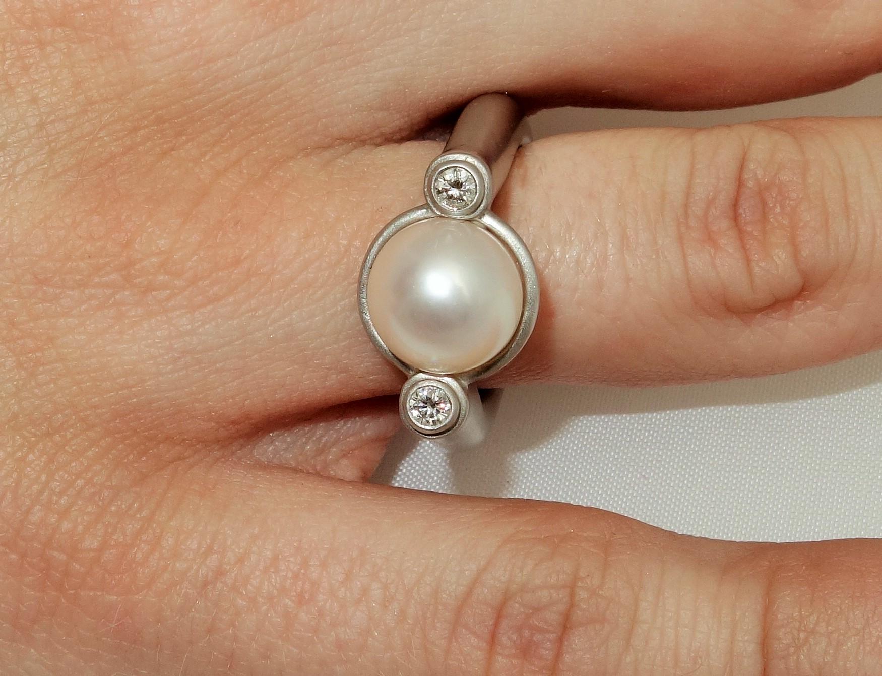 Beautiful ring featuring a 10mm Luscious Chinese freshwater Pearl held in center by a 14K pin enhanced on 2 sides with diamonds, approx. 0.13tctw; satin finish Sterling Silver Tarnish-resistant Rhodium mounting; Size 7; We offer complimentary