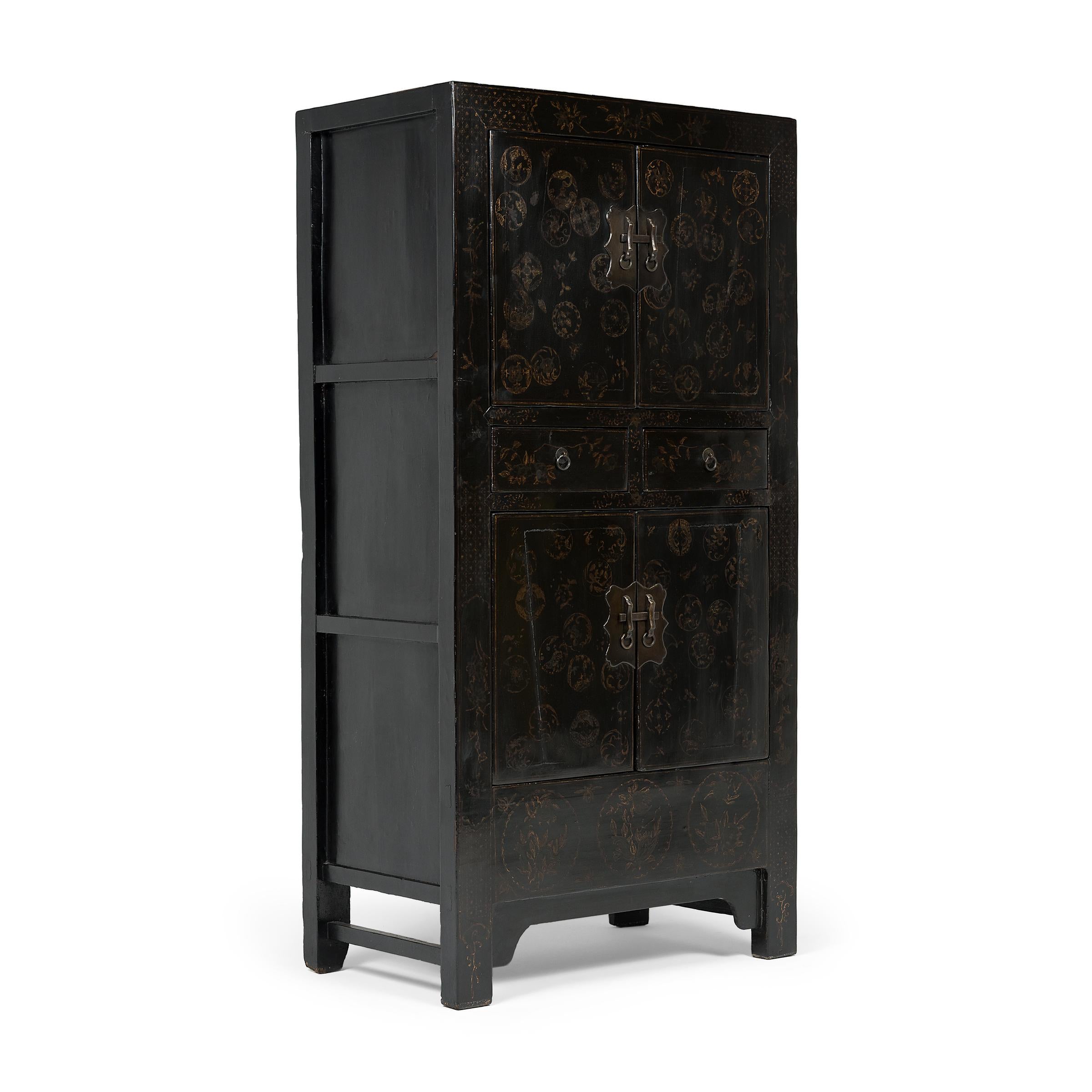 Crafted in the early 20th century, this tall cabinet has a clean-lined form designed with straight sides and square corners. The cabinet is cloaked in a glossy black lacquer finish, contrasted by hand-painted gilt decoration, now considerably faded