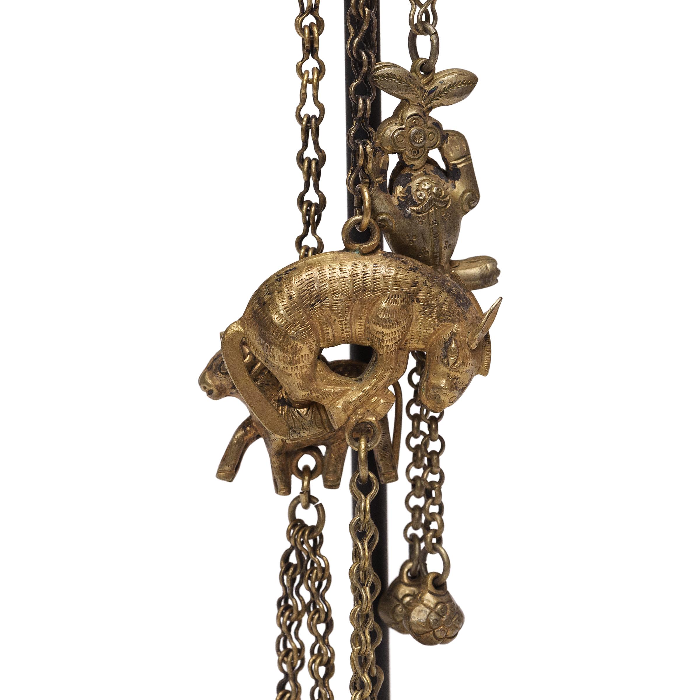 Questa curiosa catena cinese risale all'inizio del XX secolo ed è infilata in una serie di campanelle, charme e amuleti. Lavorati in lega d'argento dorata, i charms repoussé sono carichi di benedizioni e simbolismi, tra cui frutti per l'abbondanza,