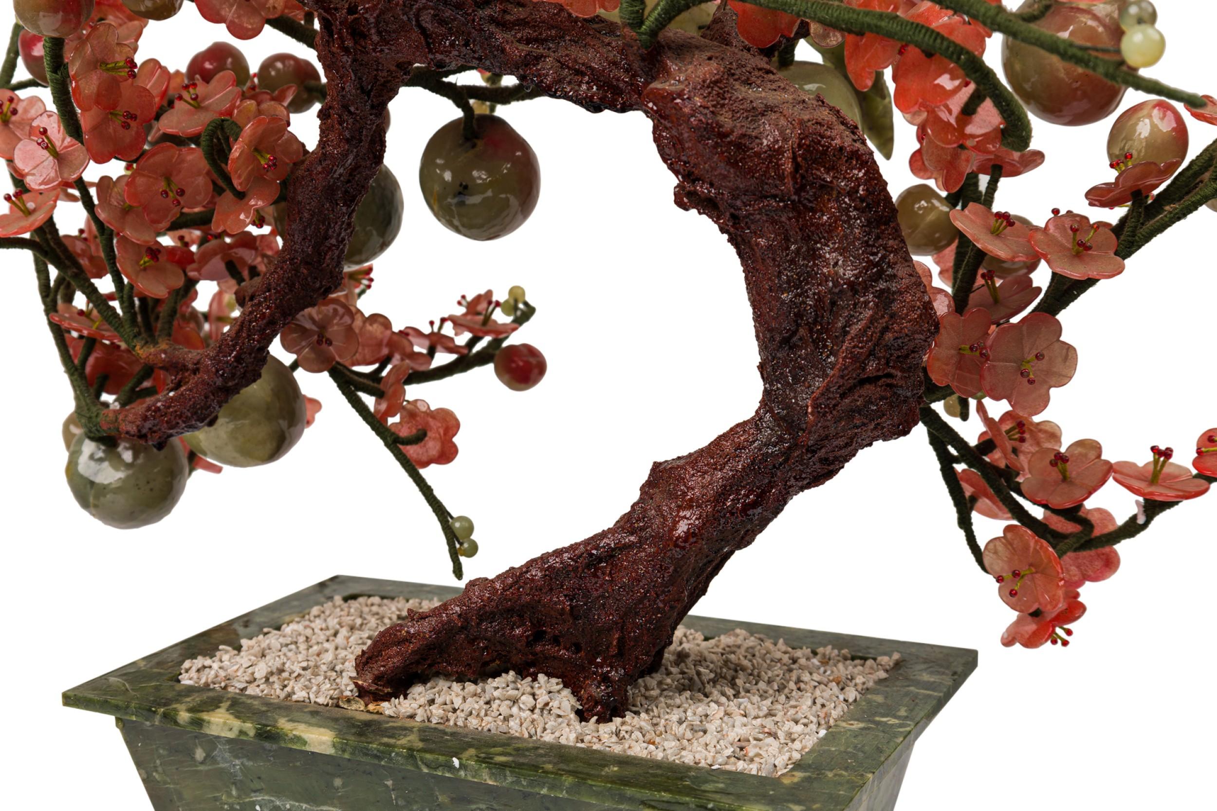 Chinese sculpture features pink glass translucent cherry blossoms and plums hanging from a lacquered tree, sprouted from a bed of faux white stones within a mottled green rectangular glass planter resembling jade.
