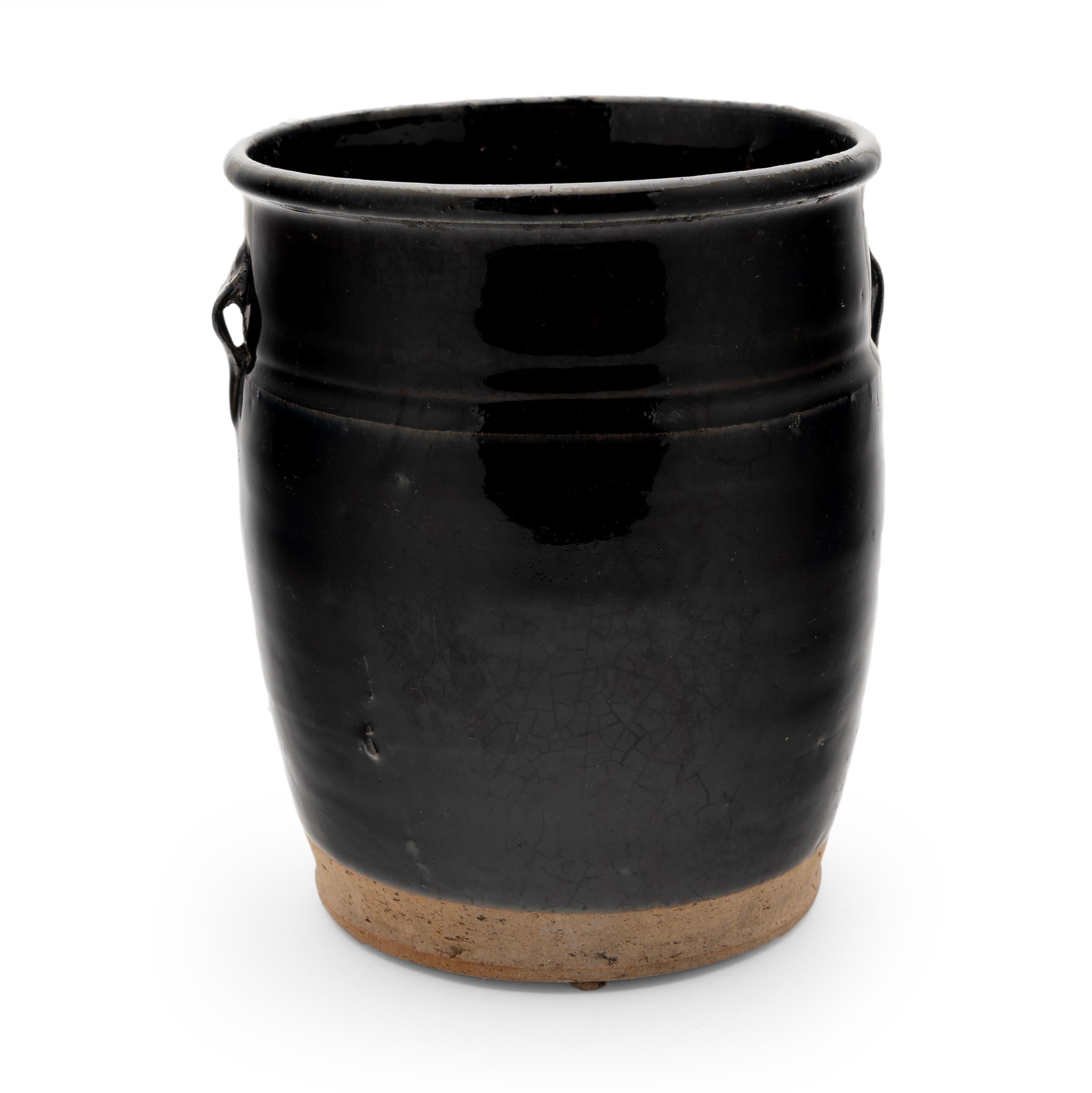 A thick black glaze softly covers the tapered form of this early 20th-century jar, once used daily in a Qing-dynasty kitchen. The petite jar is shaped with gently sloping sides and high shoulders topped by two small strap handles. The lustrous glaze