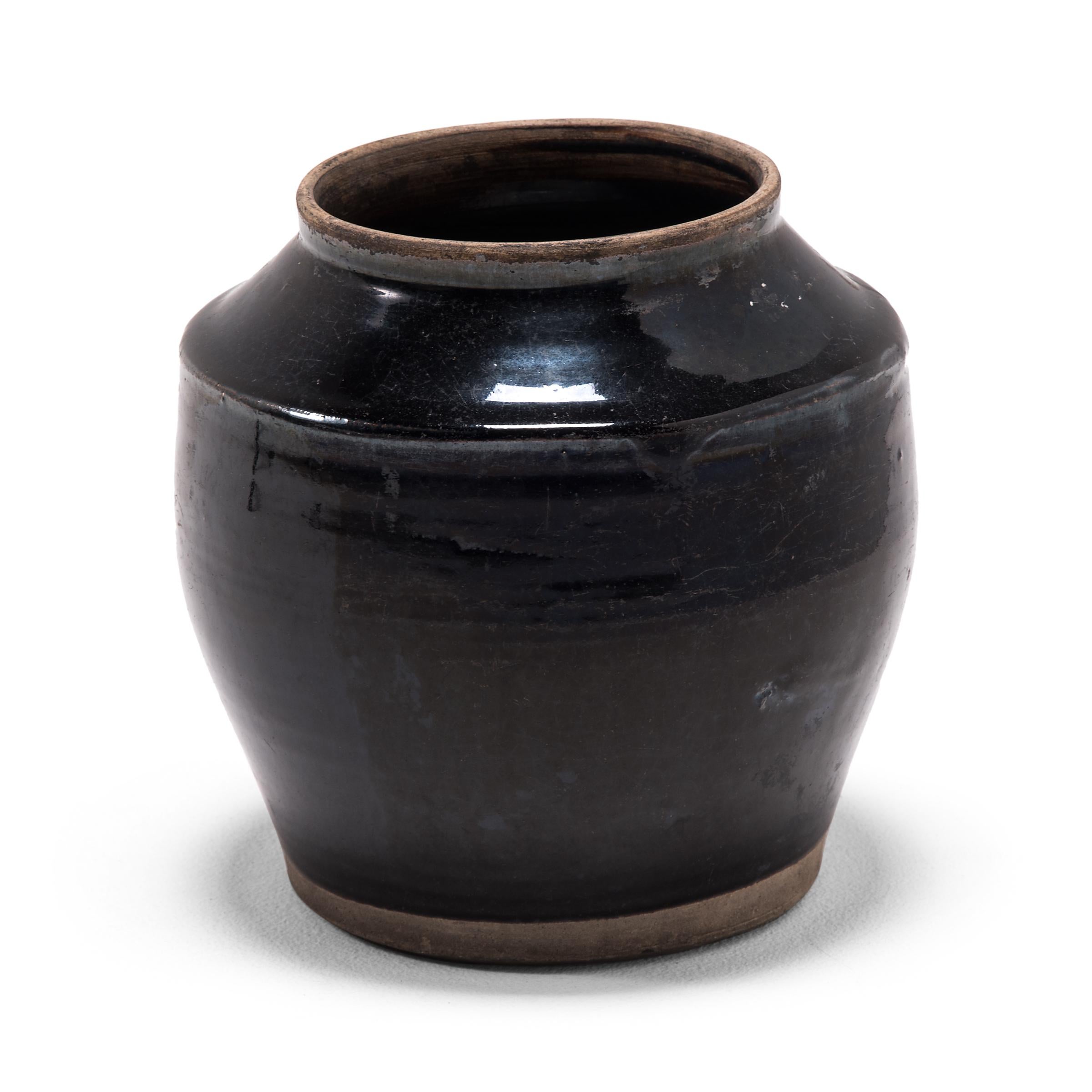 A dark glaze coats the angular body of this squat kitchen vessel, pooling in the shallow ridges along its sides with subtle iridescence. The petite early 20th century dish was once used for storing food in a Qing-dynasty kitchen, as evidenced by its