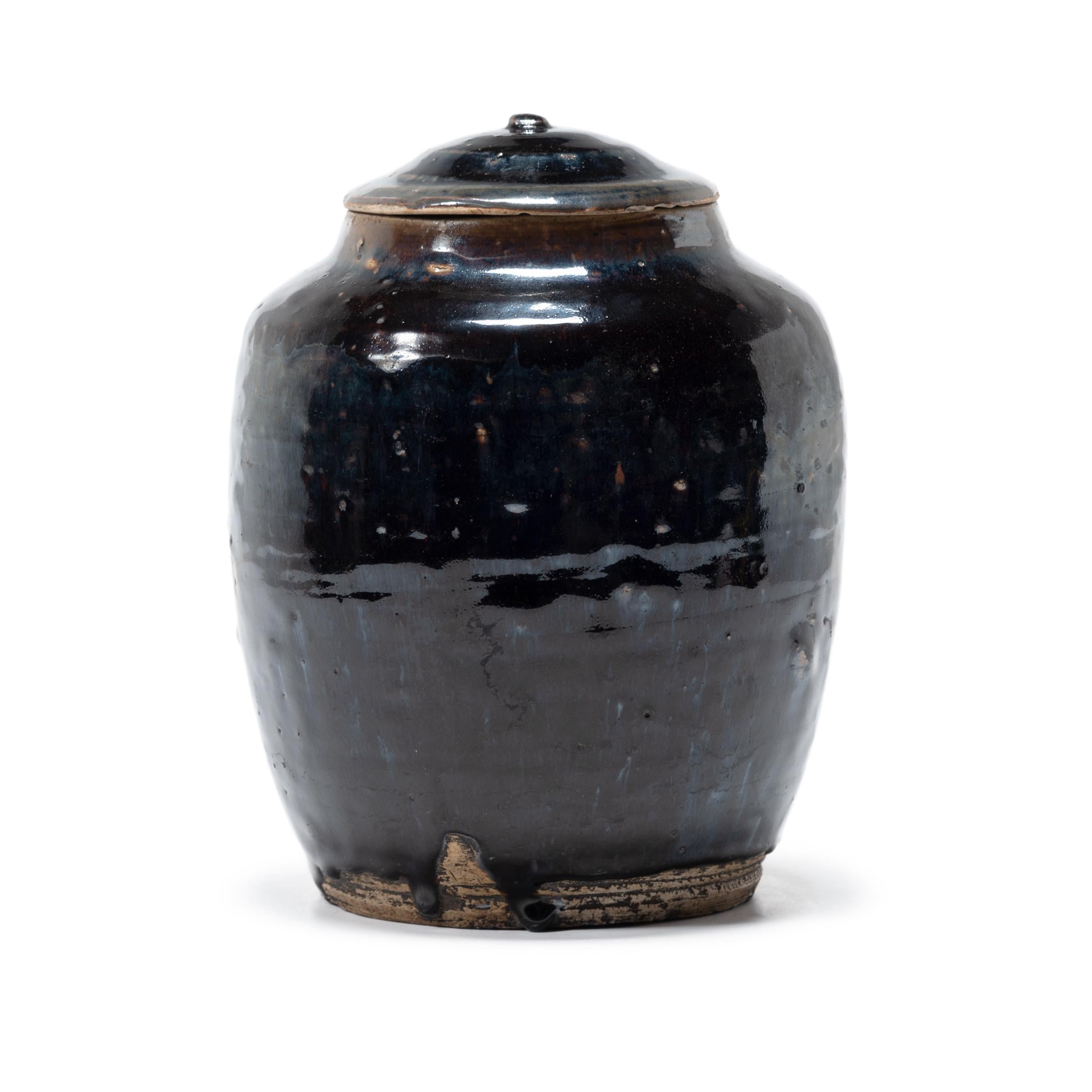 As though slicked with oil, a thick blue-black glaze spills over the high shoulders and gentle swell of this lidded kitchen vessel. The early 20th century ceramic jar was once used to store food goods in a Qing-dynasty kitchen, as evidenced by its