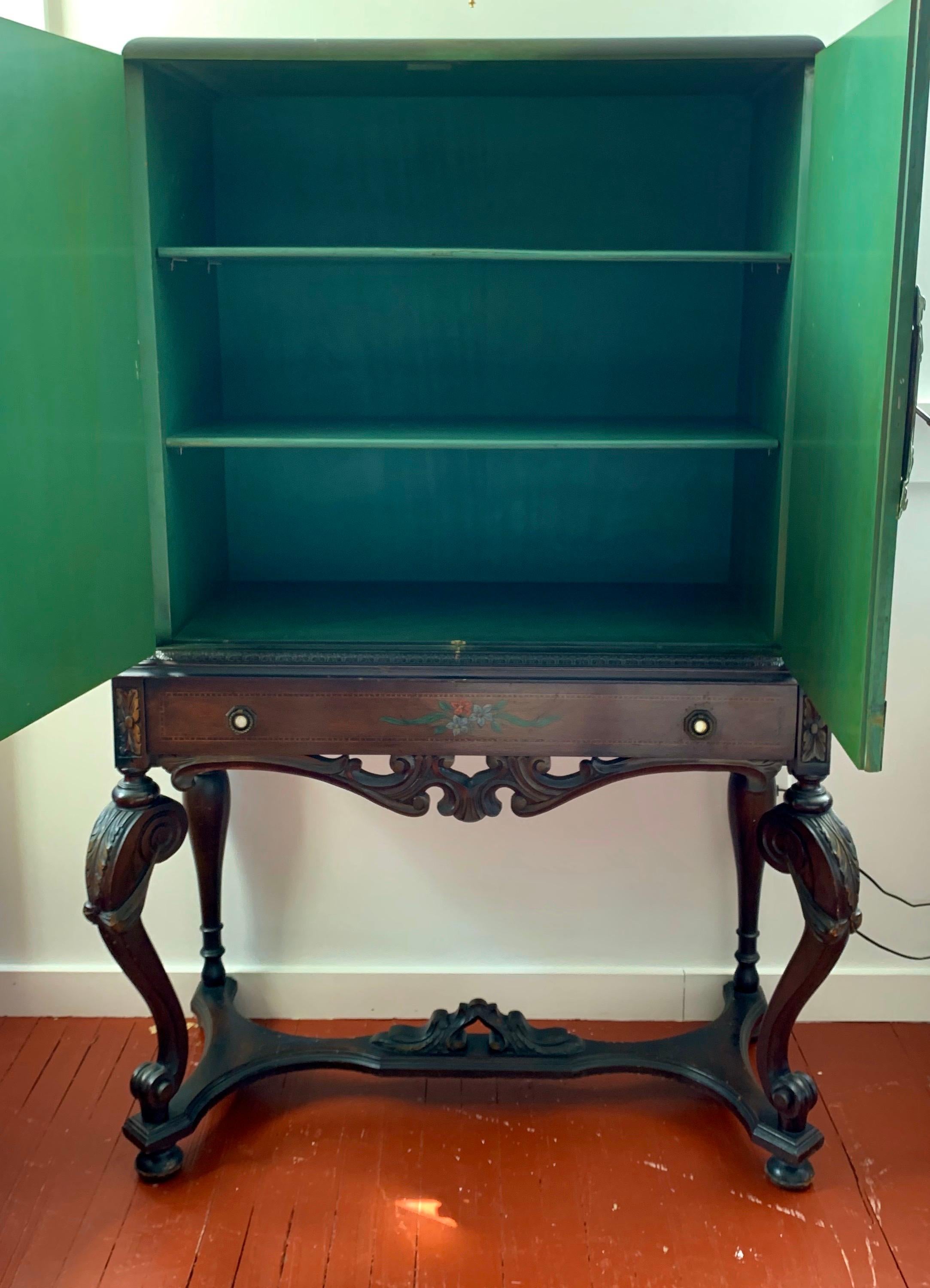 Beautiful chinoiserie cabinet on carved stand features hand painted scenery on a brilliant green background. It is painted on all three sides and has original brass filigree hardware as hinges and faceplate with key. Front doors open to shelving