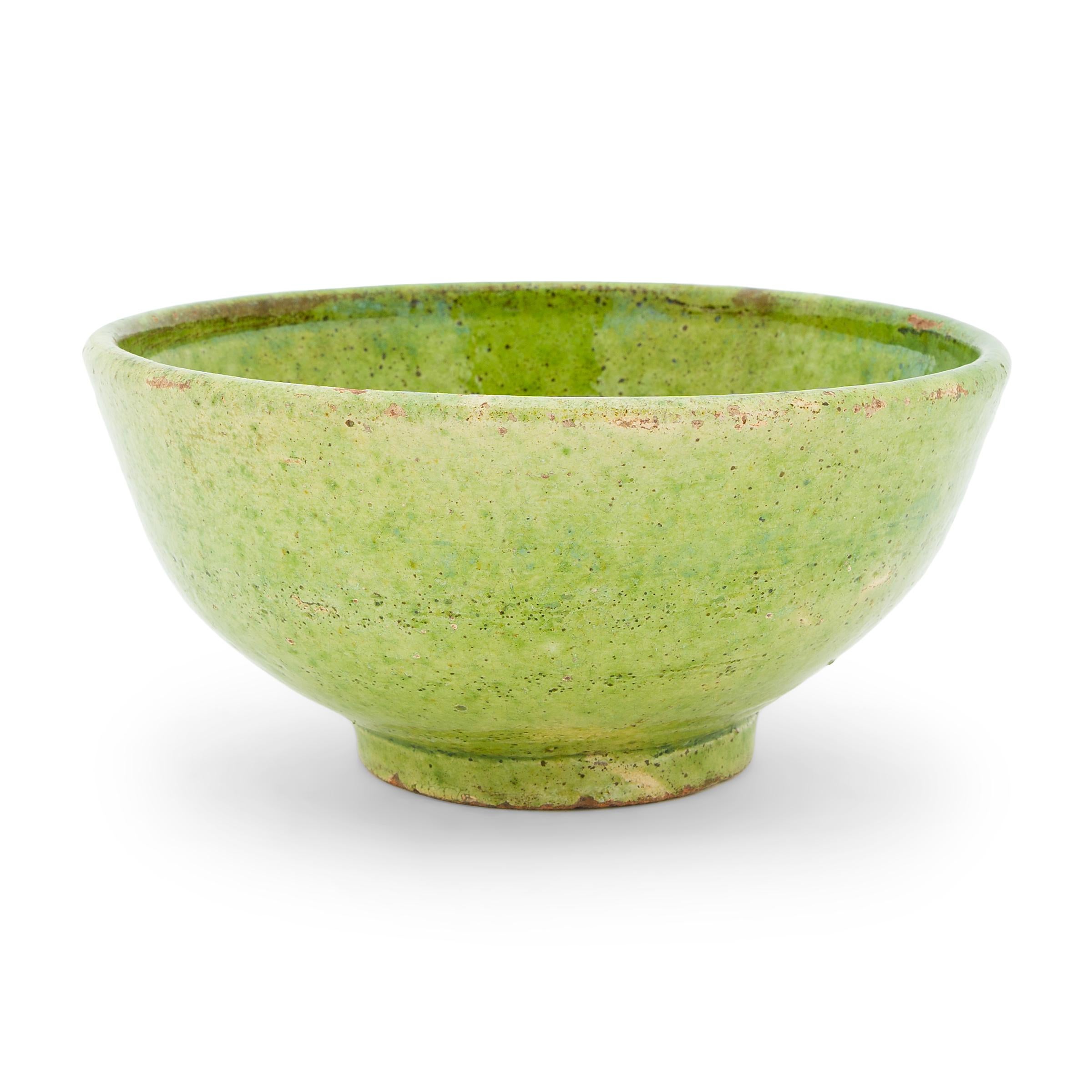 This small terracotta bowl dates to the mid-19th century and has a simple, round form with a short footed base and thin walls that flare slightly at the lip. The bowl is coated inside and out with a bright green glaze, wonderfully textured with