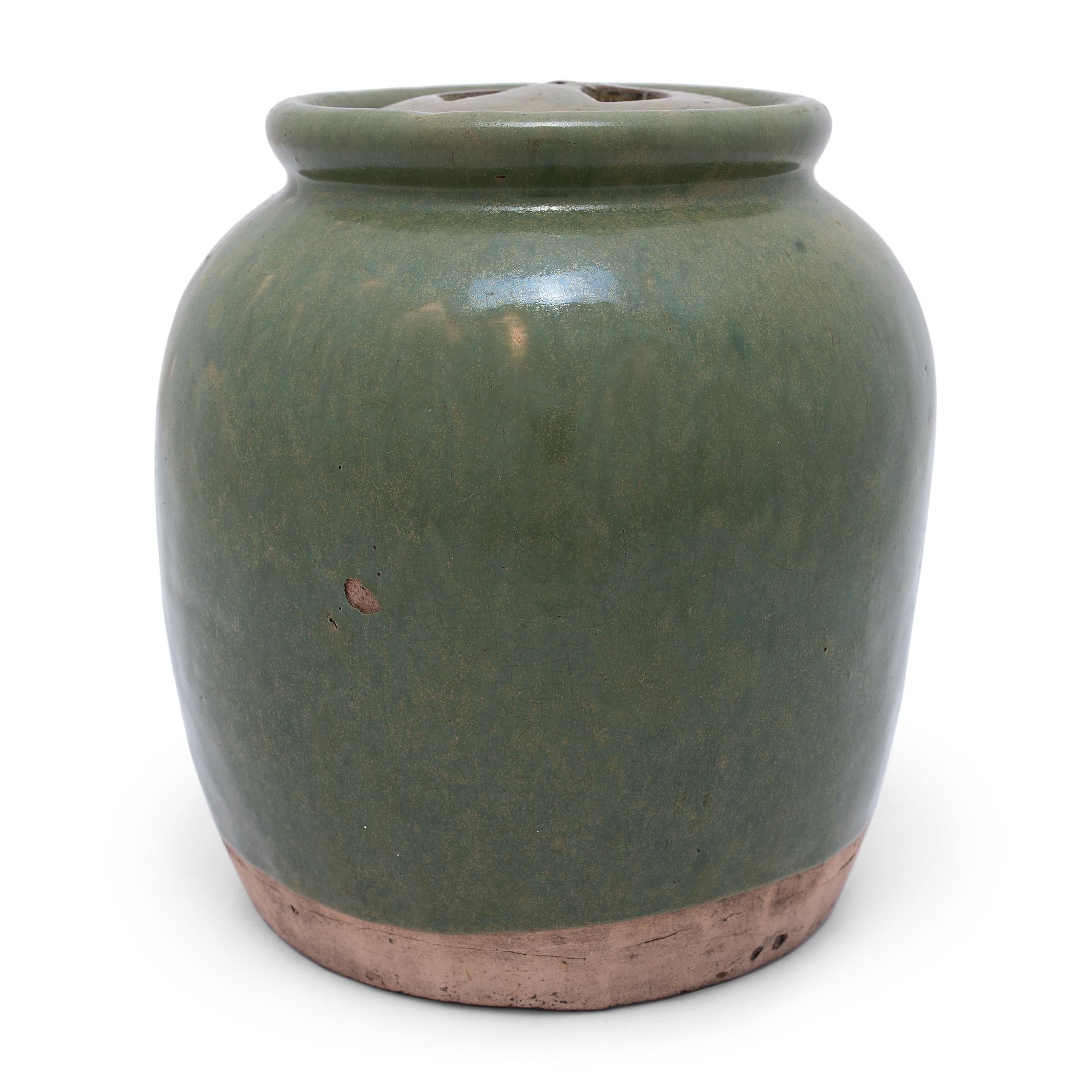 Based on forms used for pickling foods, this glazed ceramic jar has an elongated form, with high shoulders, a rounded lip, and a lid with two cutout handles. A celadon blue-green glaze sheets cloaks the exterior with beautiful irregularity, textured